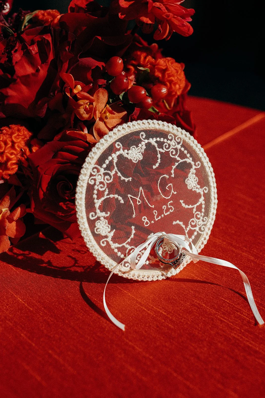 A white embroidered oval frame with monogram 'M G' and date '8.2.25', inside the frame are rings connected by a white ribbon, placed on a red surface, surrounded by red and orange flowers and berries.