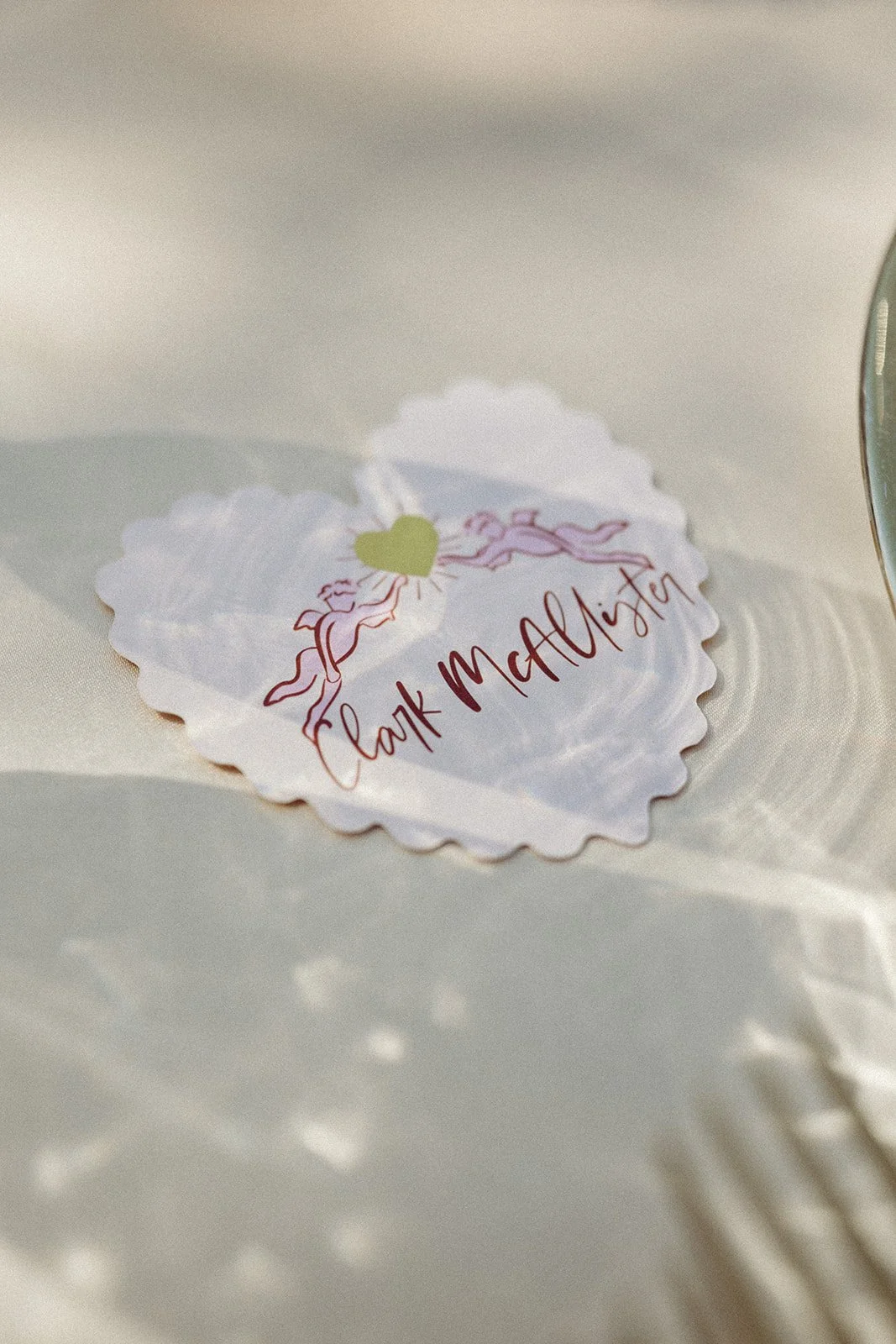 A heart-shaped paper with the name 'Clair McAlister' written on it, decorated with a pink-winged cupid illustration, placed on a white surface with shadows.