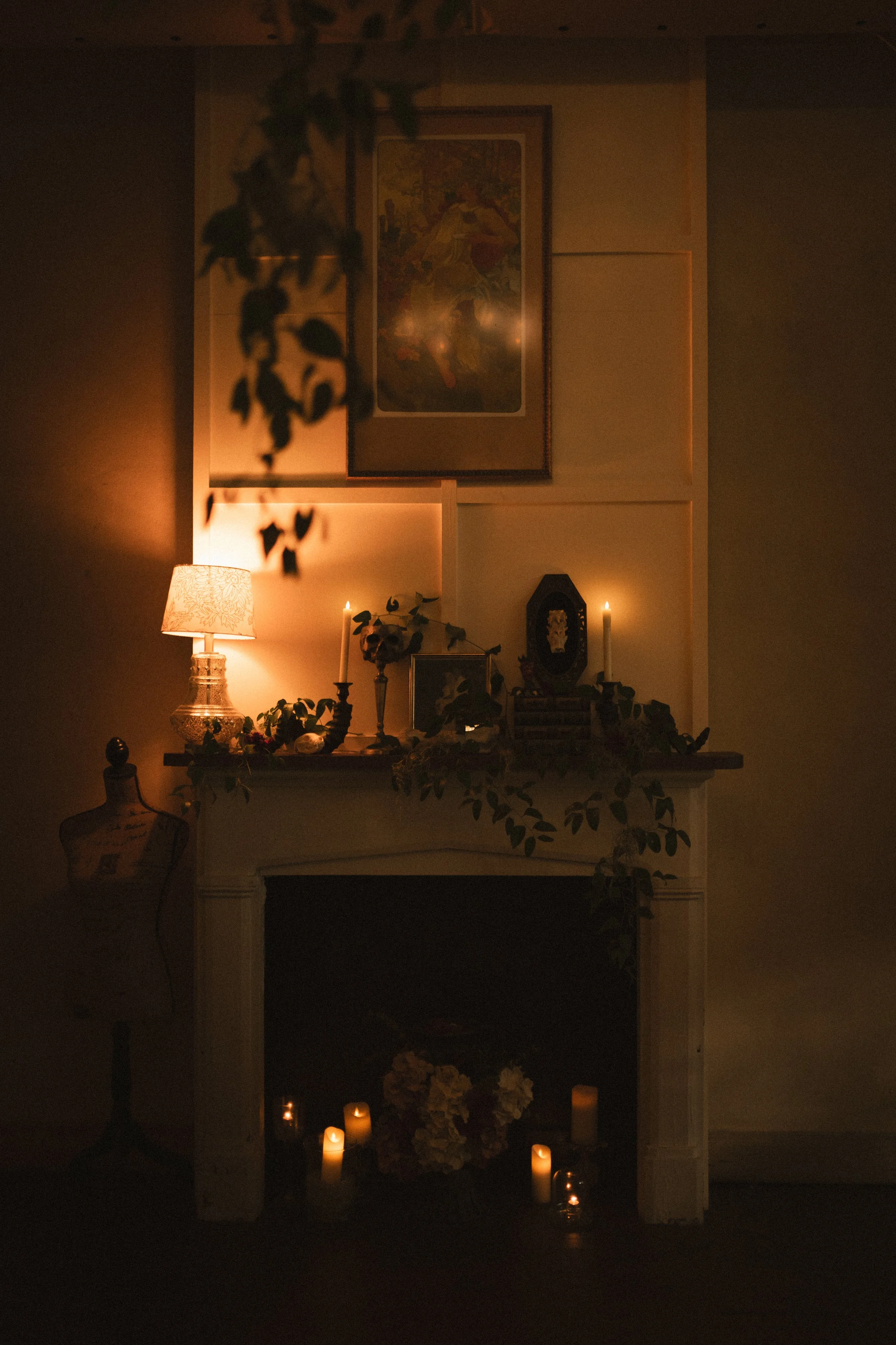 Candlelit fireplace mantel decorated with framed artwork, flowers, greenery, and a table lamp in a dimly lit room