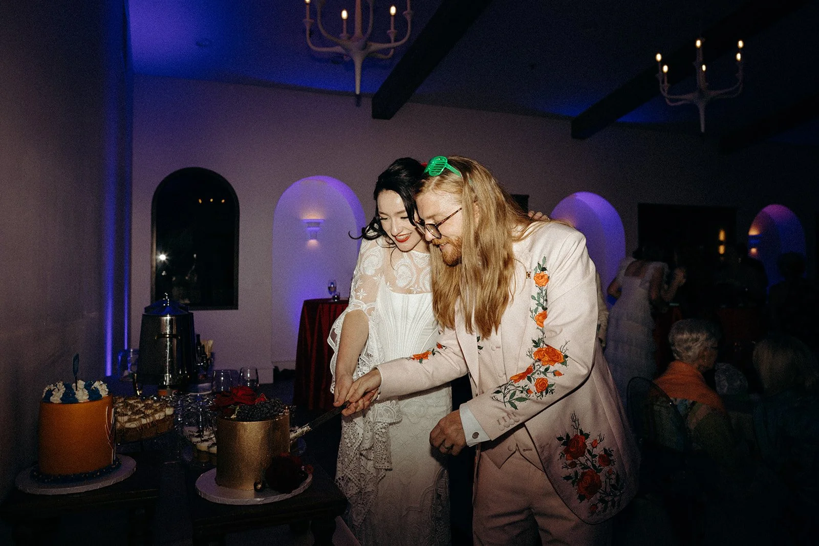 A man with long blonde hair and glasses, wearing a floral embroidered suit, is cutting a cake while a woman in a white lace dress with black hair smiles beside him. They are at a celebration, with cakes on the table and other guests in the background