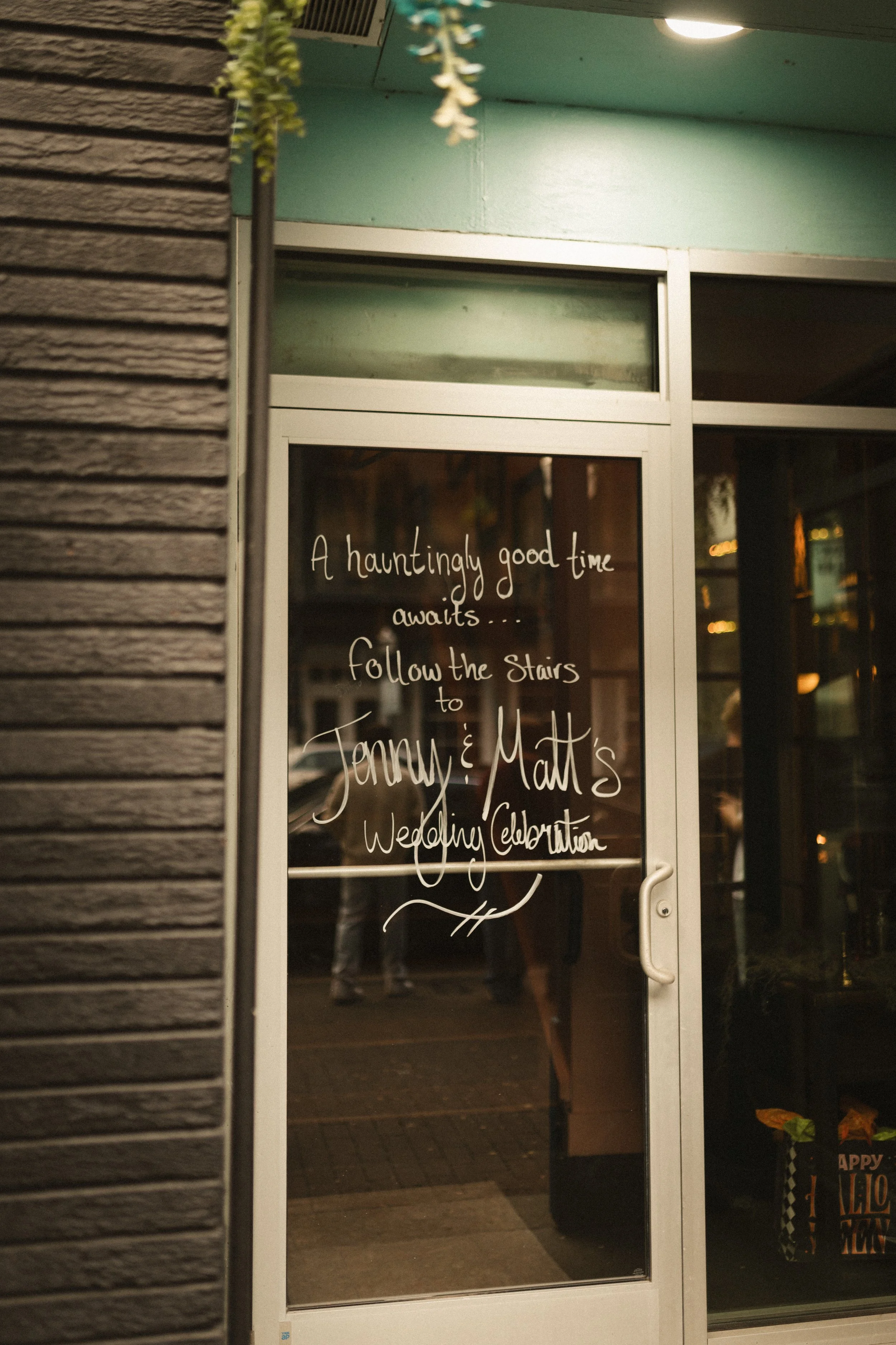 Halloween-themed storefront door with handwritten message reading “A hauntingly good time awaits… Follow the stairs to Jenny & Matt's Wedding Celebration.”