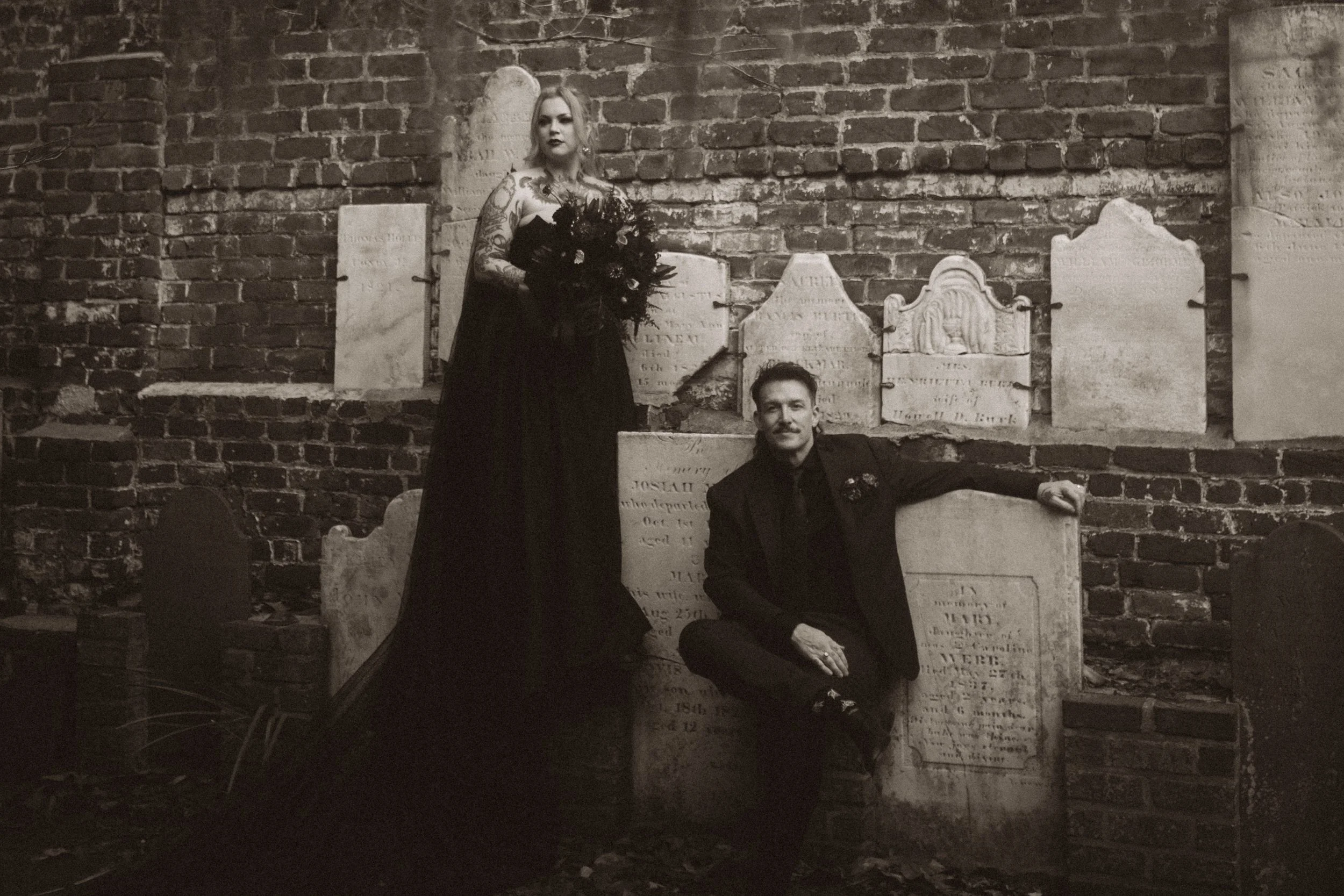 Black-and-white portrait of a tattooed woman in a long black dress and a seated man surrounded by antique gravestones against a brick wall