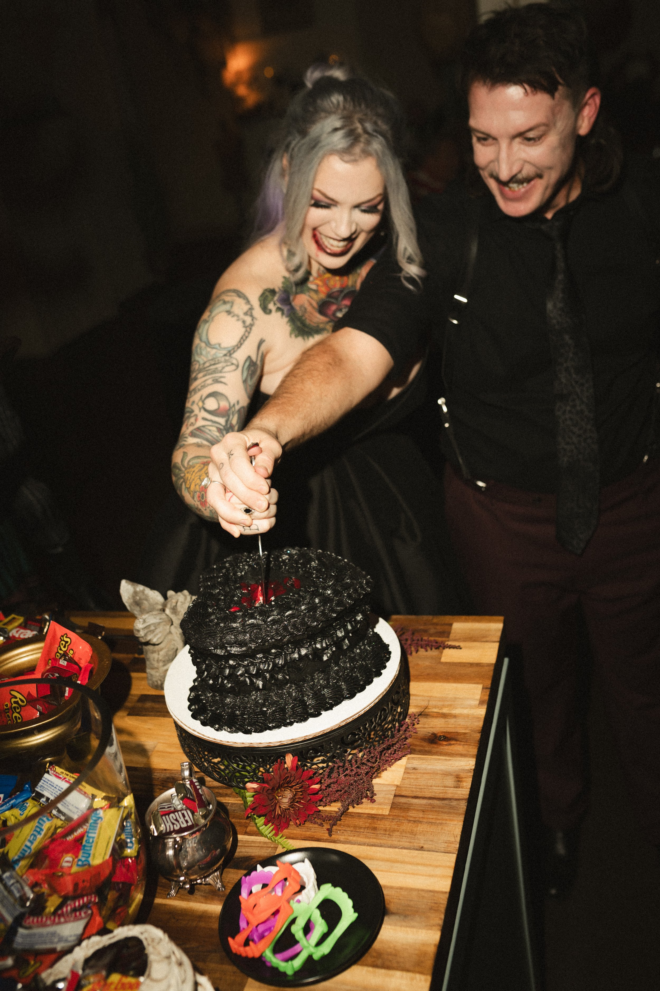 Tattooed woman and man laughing beside a tall black cake at a Halloween-themed party