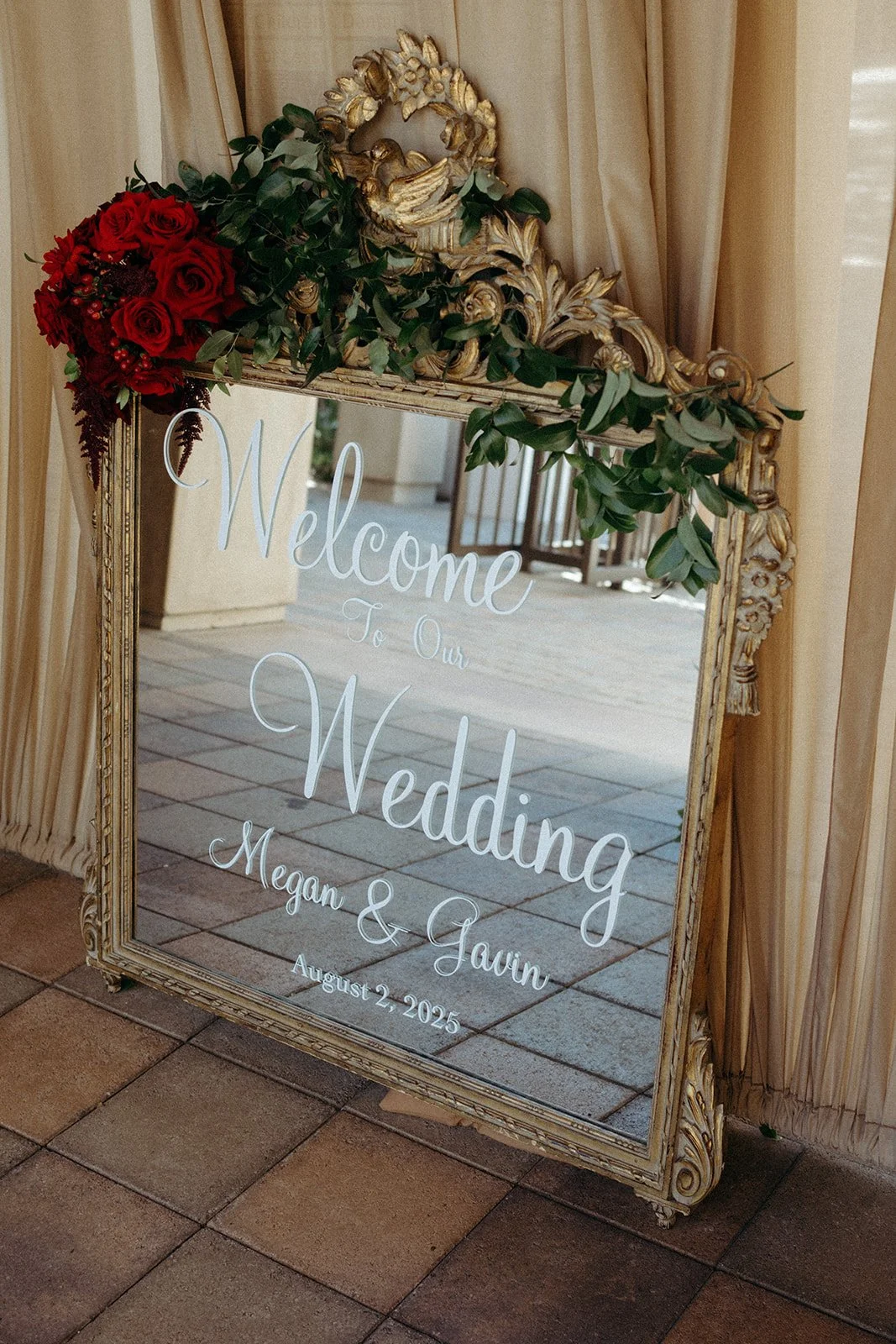 Decorative mirror with gold ornate frame, adorned with red roses and greenery, displaying a welcome message for a wedding of Megan and Gavin on August 2, 2025.