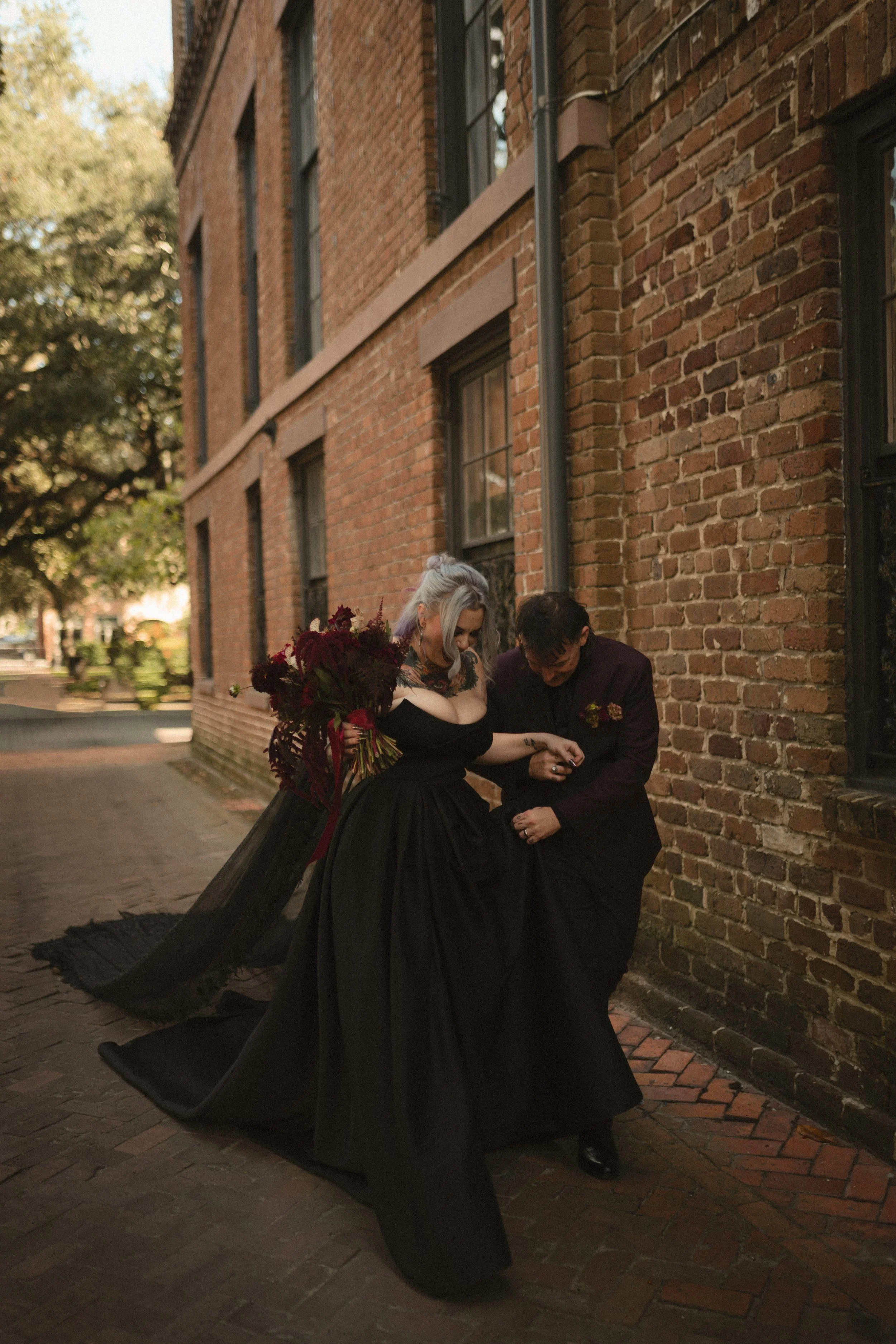 Two people in black outfits pose with a dark floral bouquet against a brick building.