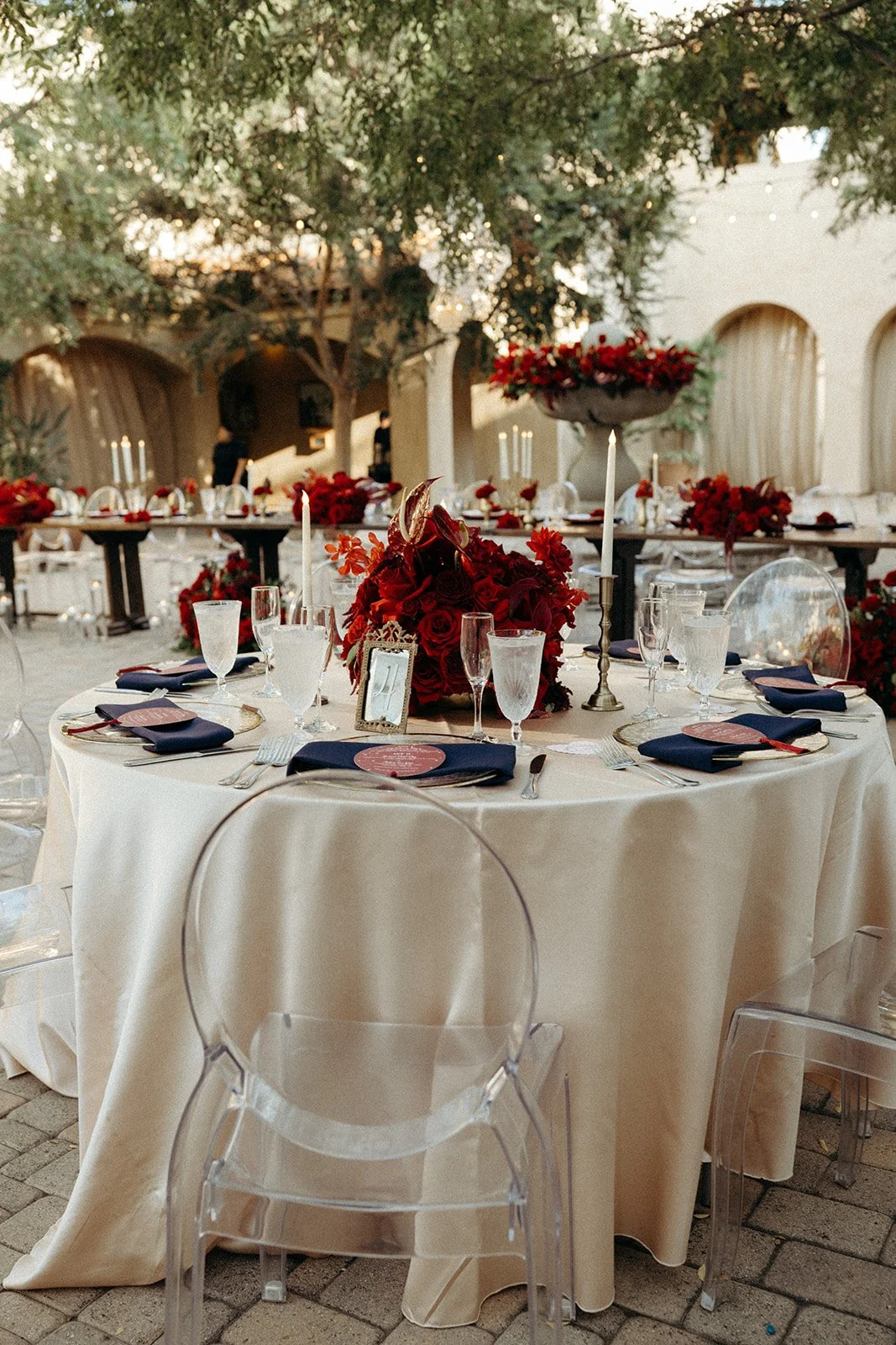 Elegant outdoor wedding reception with round table set with navy blue napkins, glassware, and gold cutlery, featuring a centerpiece of red roses and foliage, surrounded by transparent ghost chairs, under large trees with string lights overhead.