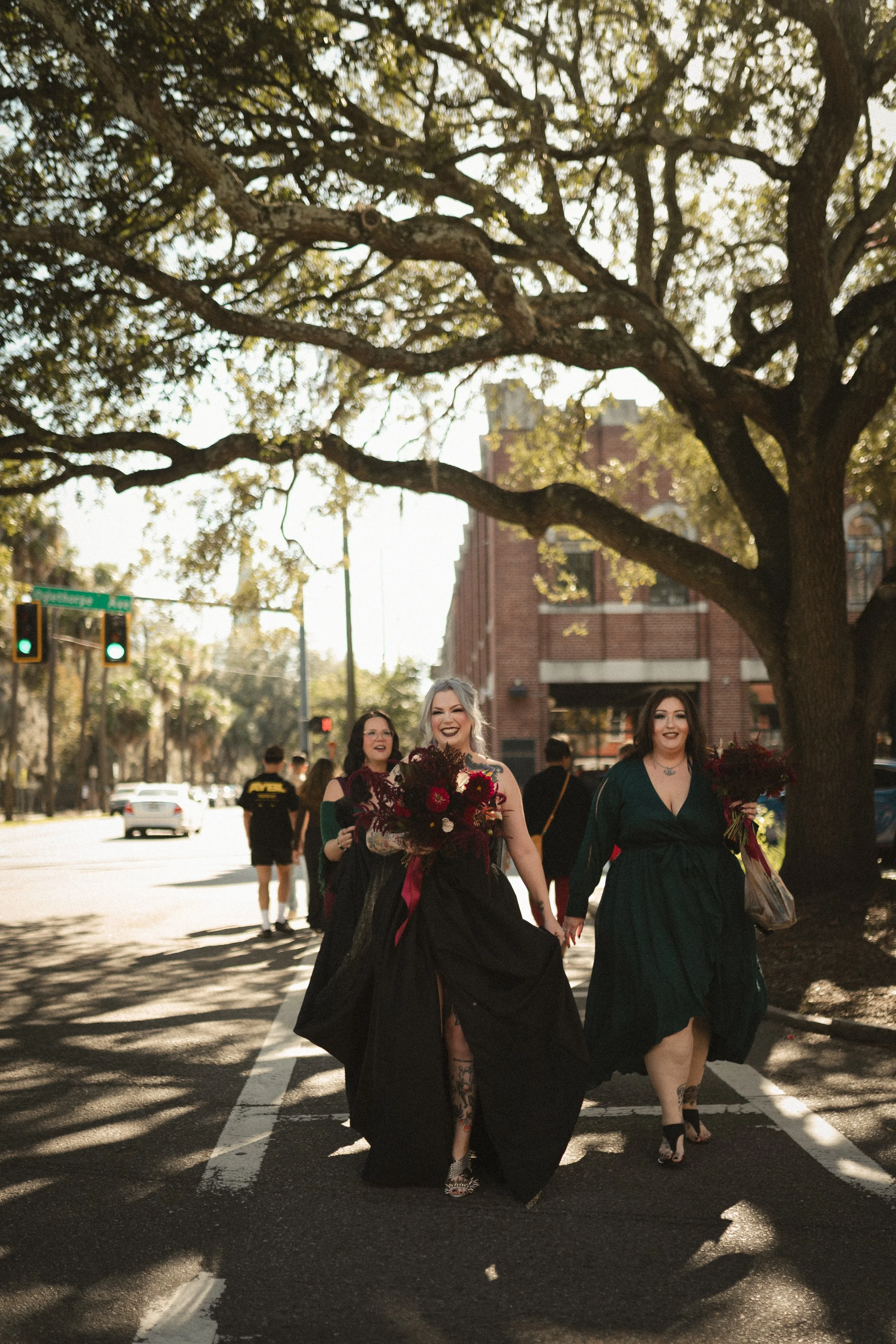 A smiling bride and bridesmaids carrying bouquets walk across a city street beneath large oak trees.