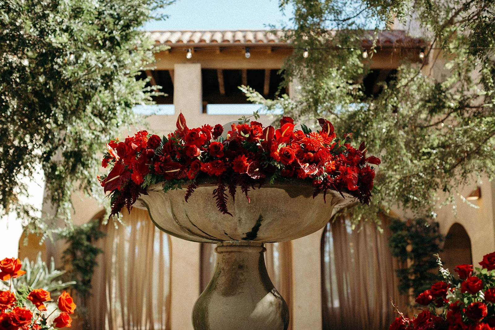 A large urn filled with red flowers, set outdoors with trees and a building in the background.