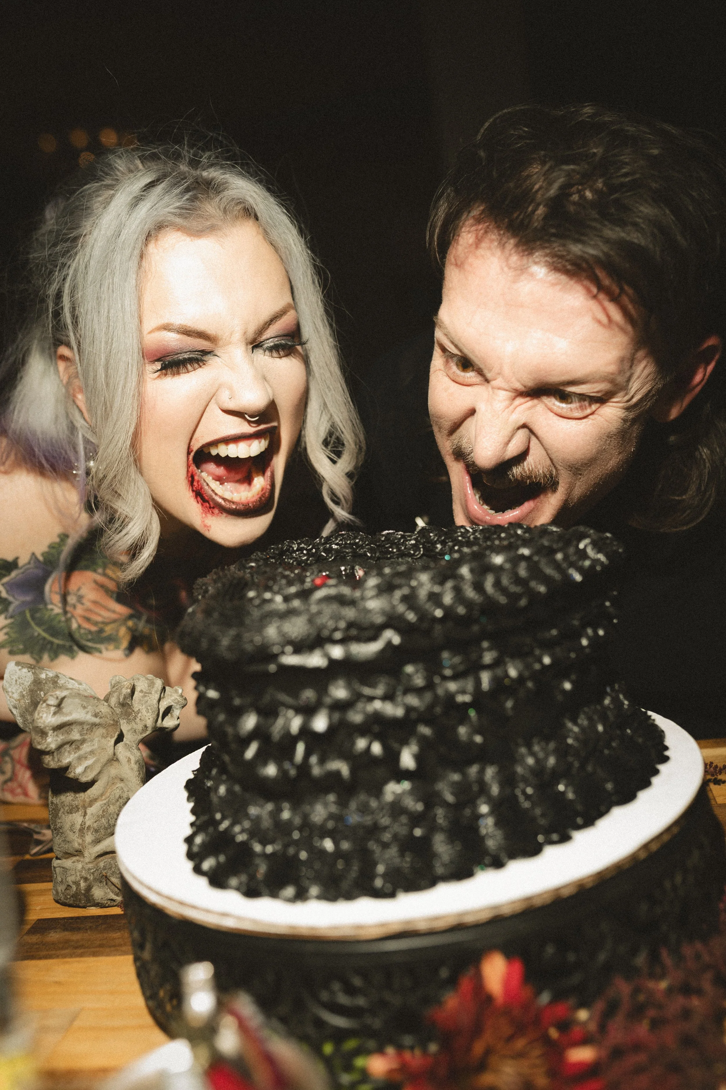 Two people in Halloween makeup laughing beside a black glitter-covered cake on a white plate.