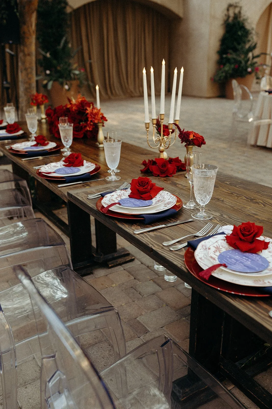 A decorated dinner table set for a special occasion with floral and candle centerpieces, elegant plates, silverware, and glasses, featuring red roses and a candelabra with lit candles.