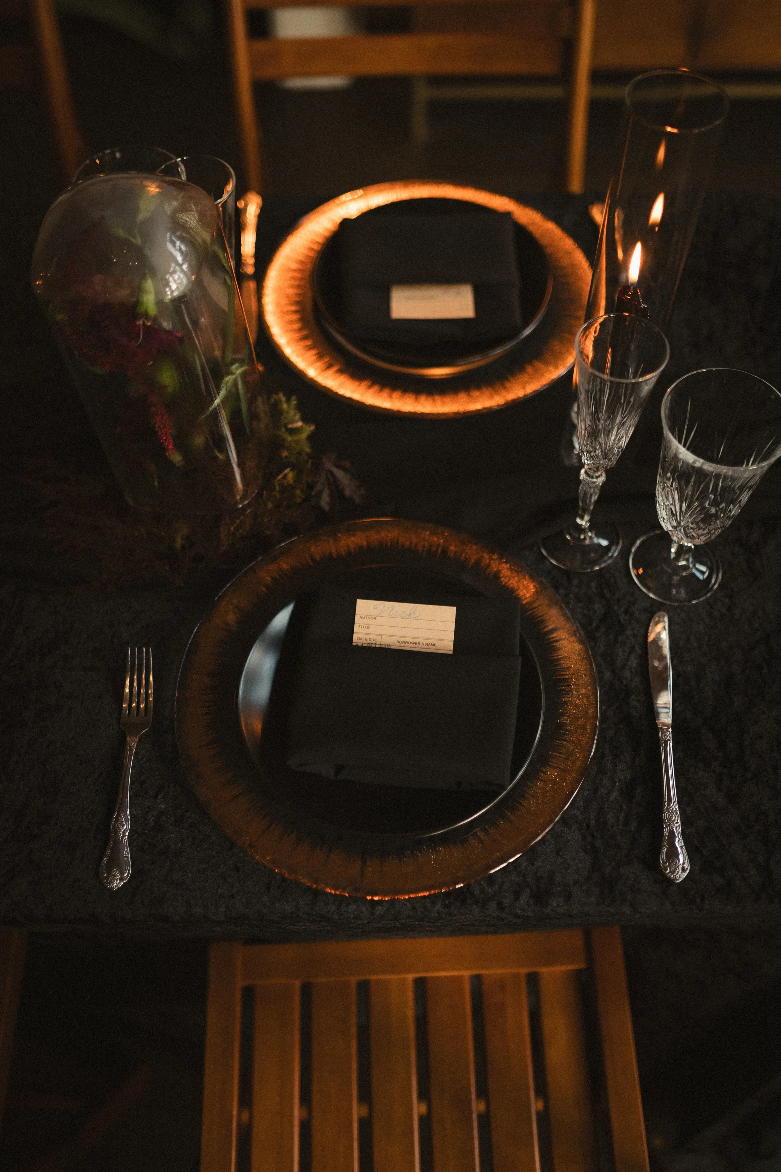 Moody Halloween dinner table with black plates, amber chargers, crystal glasses, candles, flowers, and a vintage library card place setting