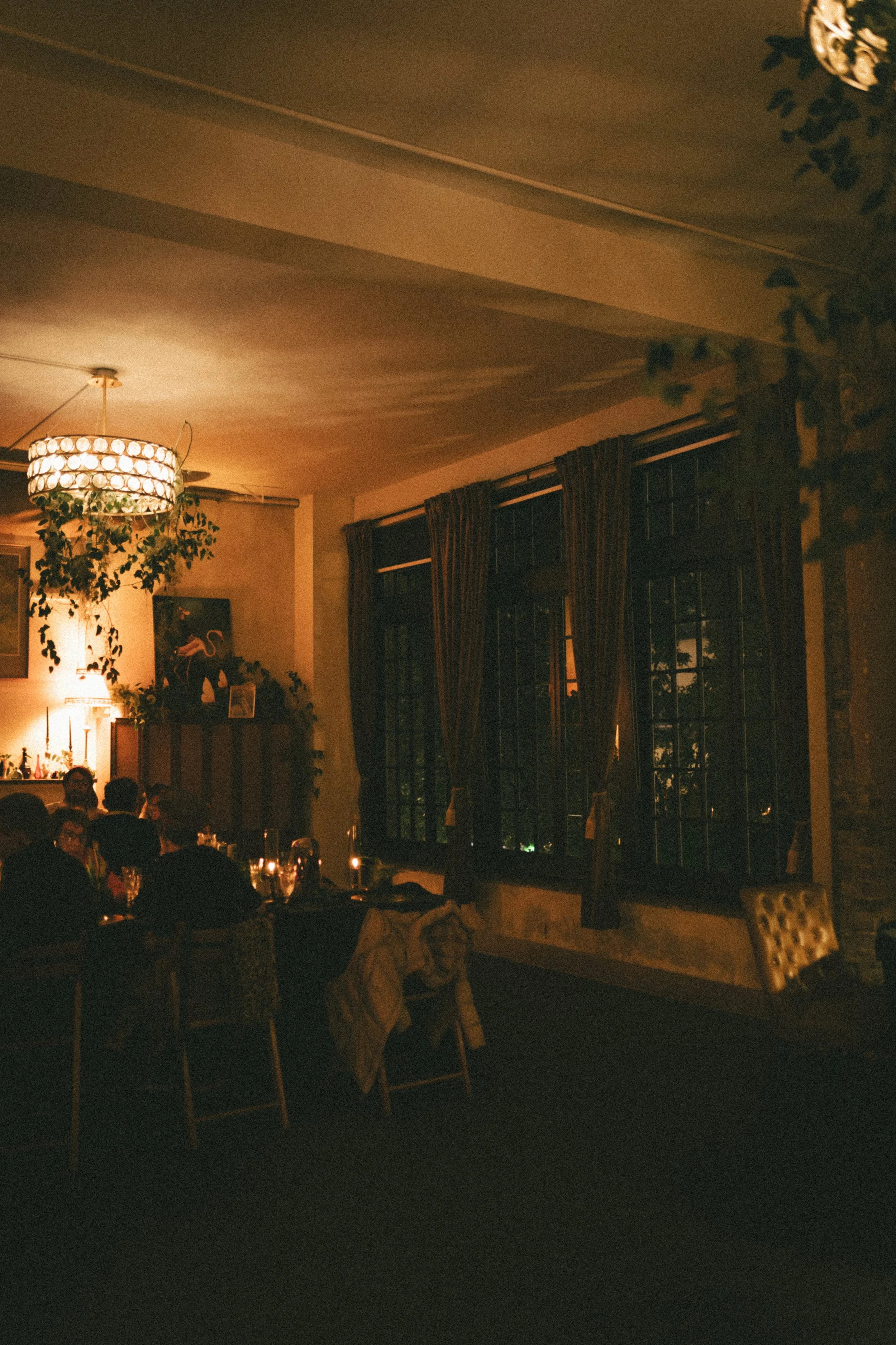 Dimly lit restaurant interior with a large crystal chandelier, hanging plants, dark curtains, and seated diners.