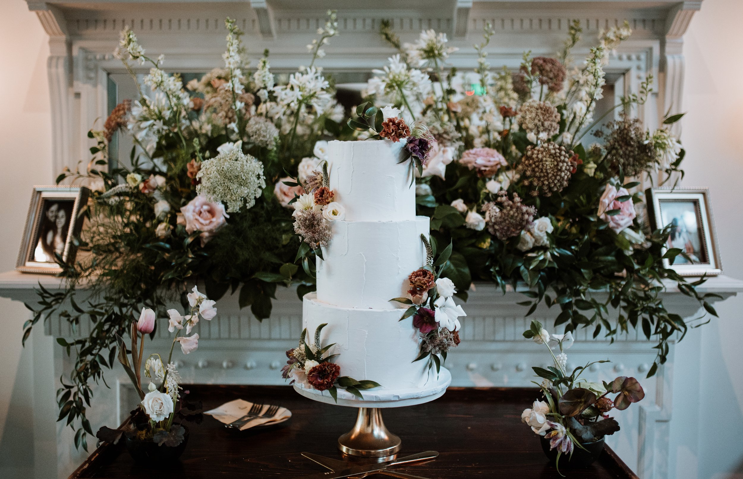 A white, three-tiered wedding cake decorated with flowers and greenery, placed on a gold cake stand. The cake is surrounded by a large floral arrangement with various flowers and framed photographs in the background.