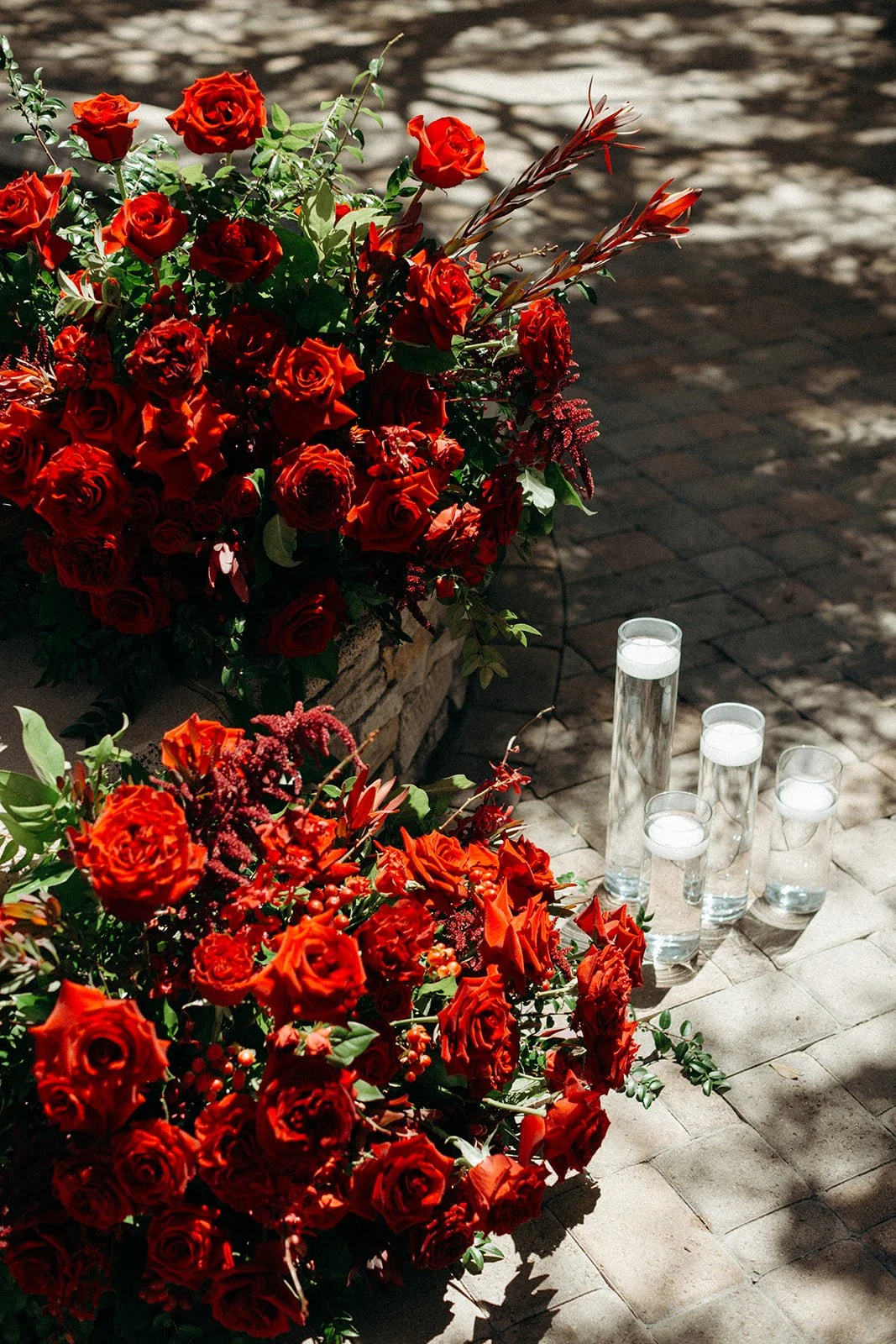 A display of two large arrangements of red roses and other red flowers, with four tall glasses of water positioned on the ground near the flowers, casting shadows on the brick pavement.