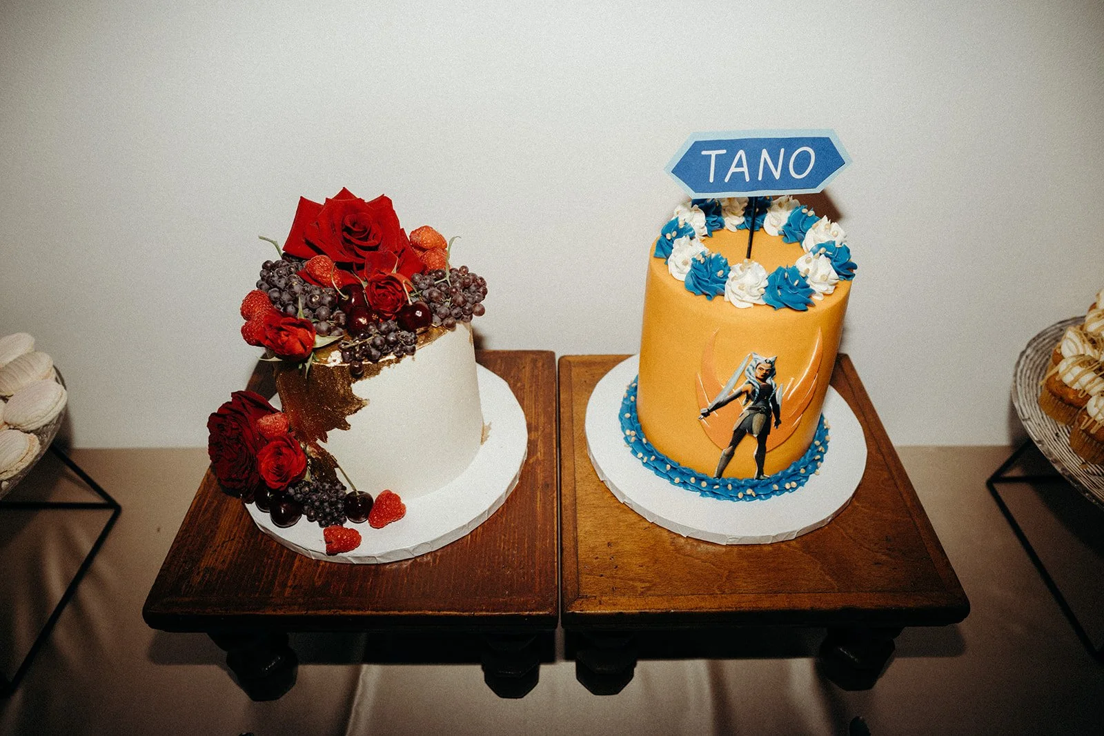 Decorative cakes with themed designs, one with fresh fruit and roses, the other with a 'TANO' sign and a Star Wars character figure, placed on wooden tables against a plain wall.