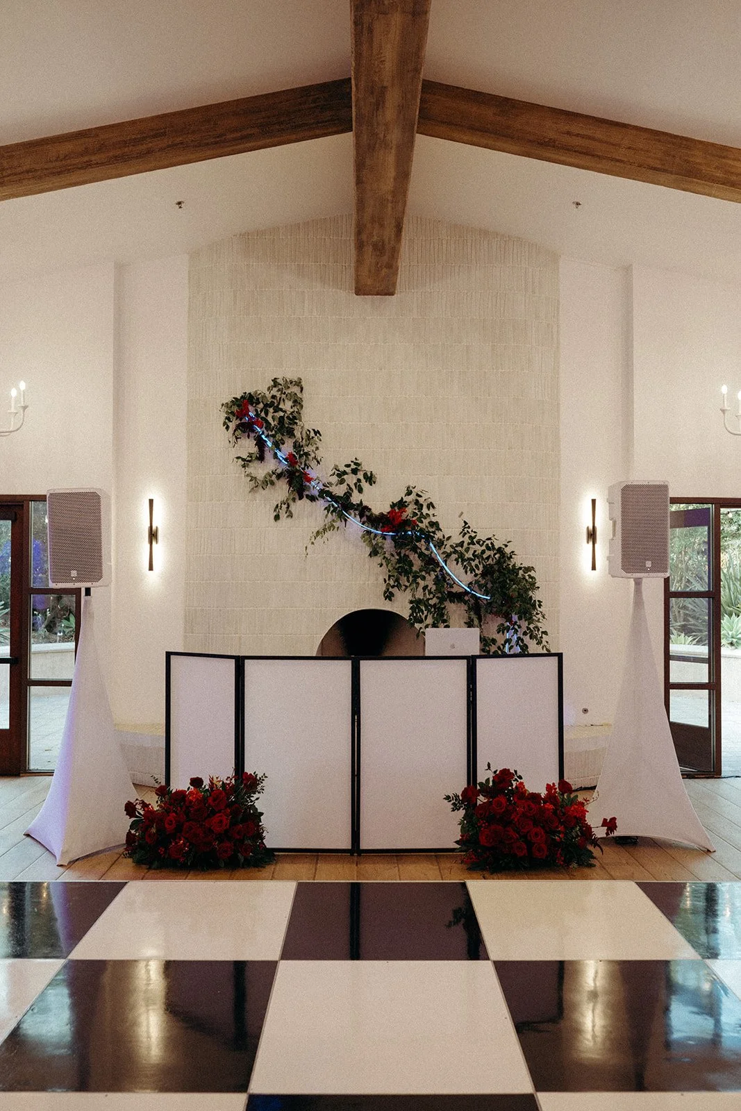 Wedding or event setup with a white partition, floral arrangements with red flowers, green foliage, and decorative lighting in a room with wood beams, windows, and black-and-white checkered floors.