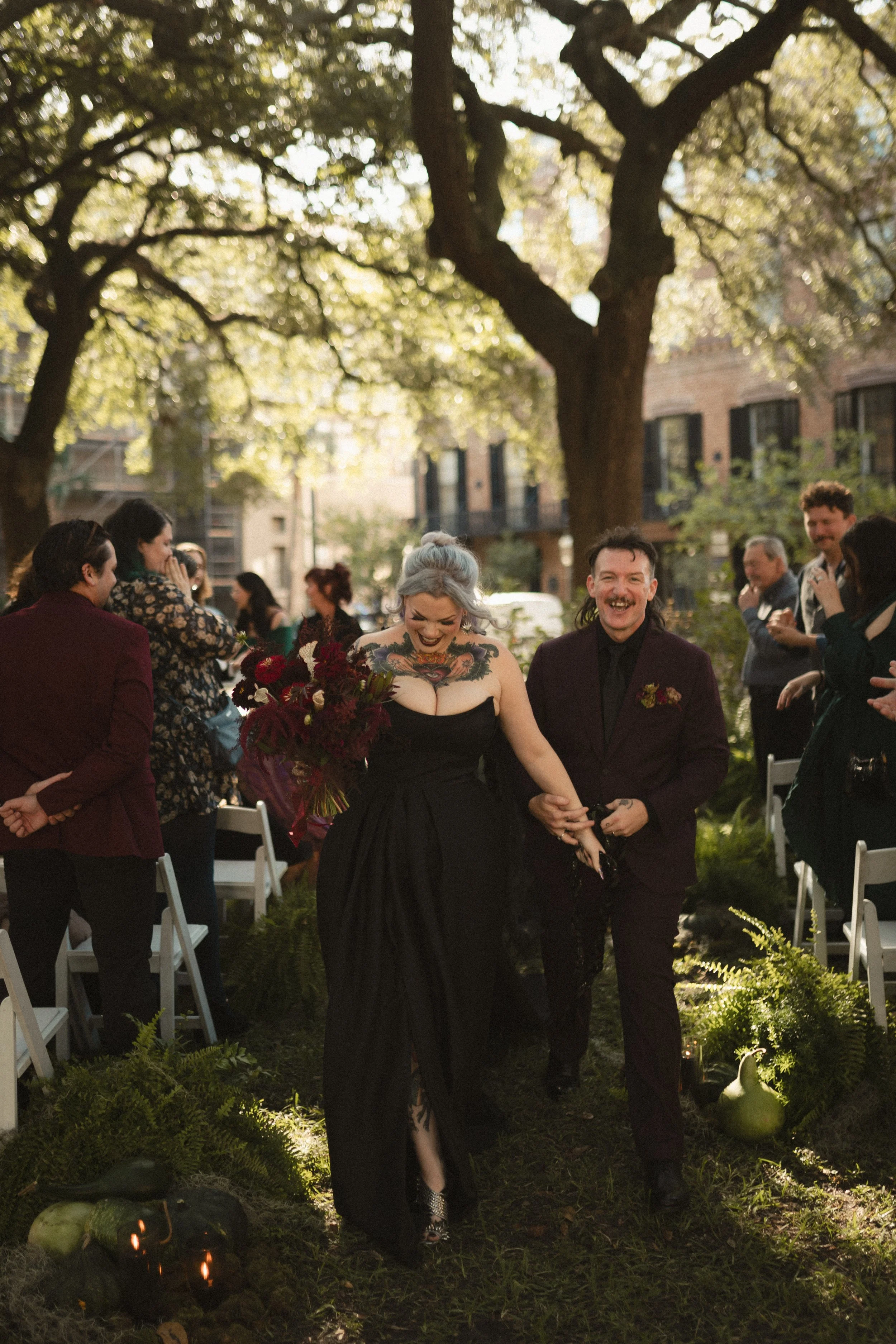 Wedding guests celebrate outdoors as a tattooed bride in a black dress carries a bouquet beside her smiling partner under sunlit trees.