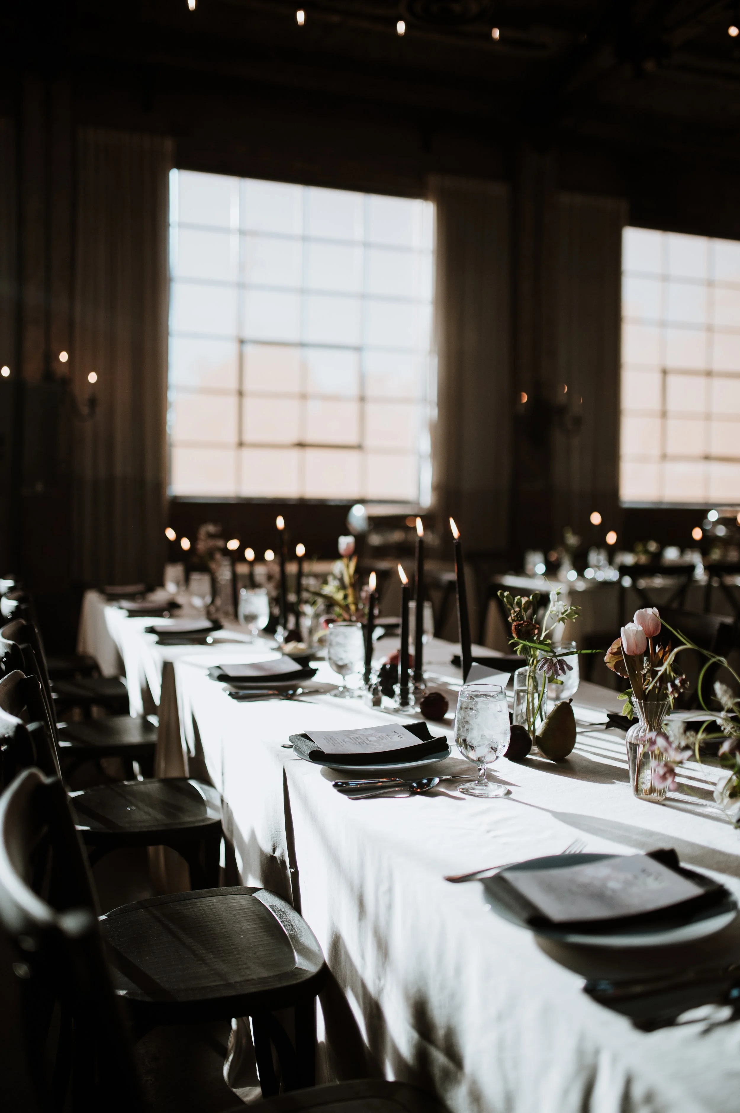 Elegant banquet table set with black candleholders, flowers in vases, and place settings, in a dimly lit room with large windows and curtains.