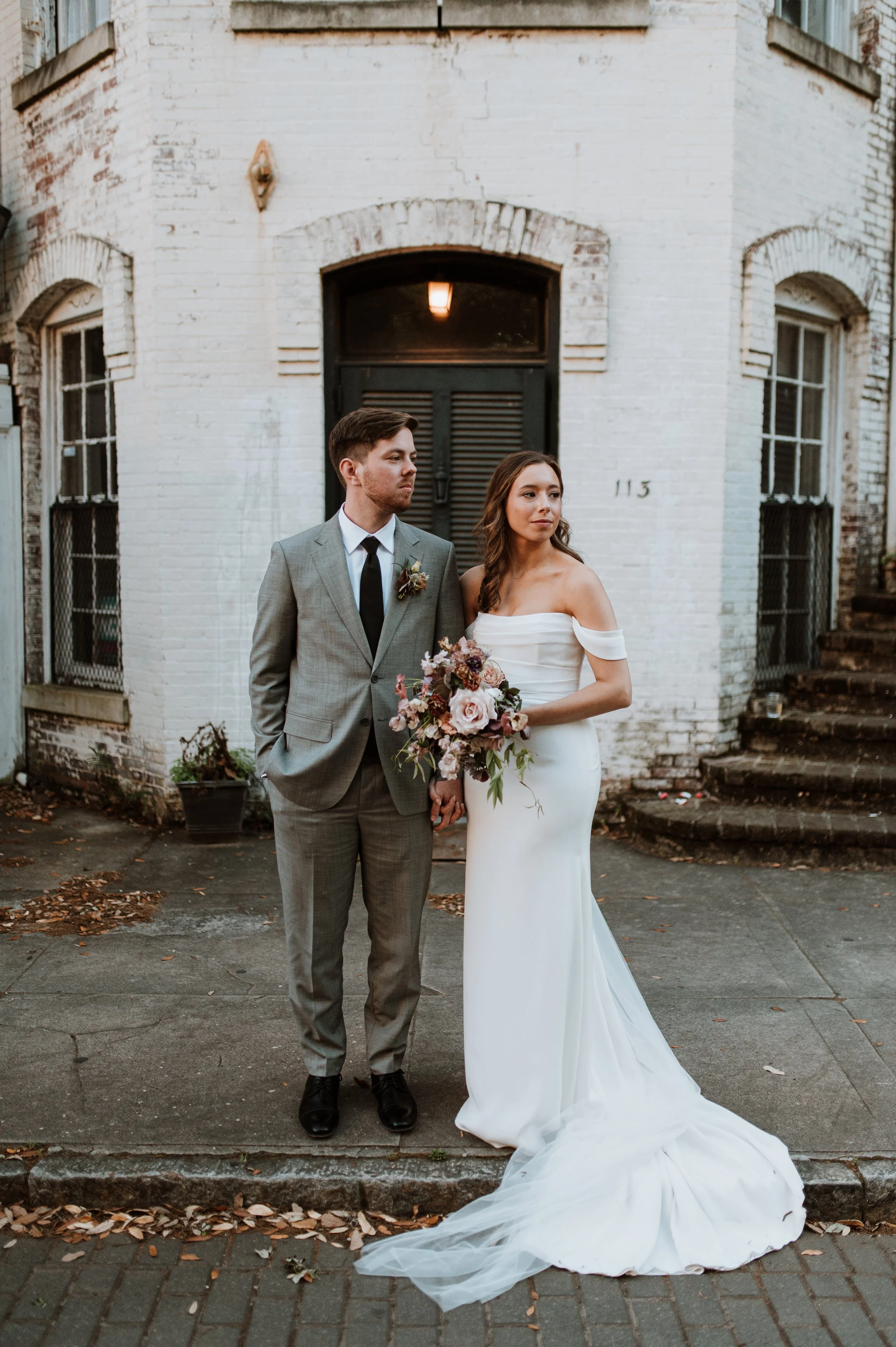 A bride and groom holding hands stand outside in front of a white brick building with arched windows and a black door. The bride is wearing a white off-the-shoulder wedding gown and holding a bouquet, while the groom is dressed in a gray suit with a black tie and boutonniere. Fallen leaves and steps are visible on the ground.