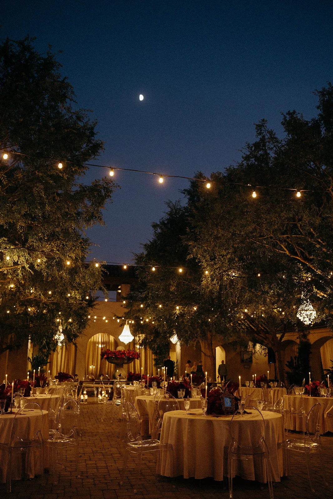 An outdoor evening event with round tables covered in cream tablecloths, decorated with floral centerpieces and lit candles. String lights hang above, illuminating the area amid trees. People are seen in the background near a stage with curtains and