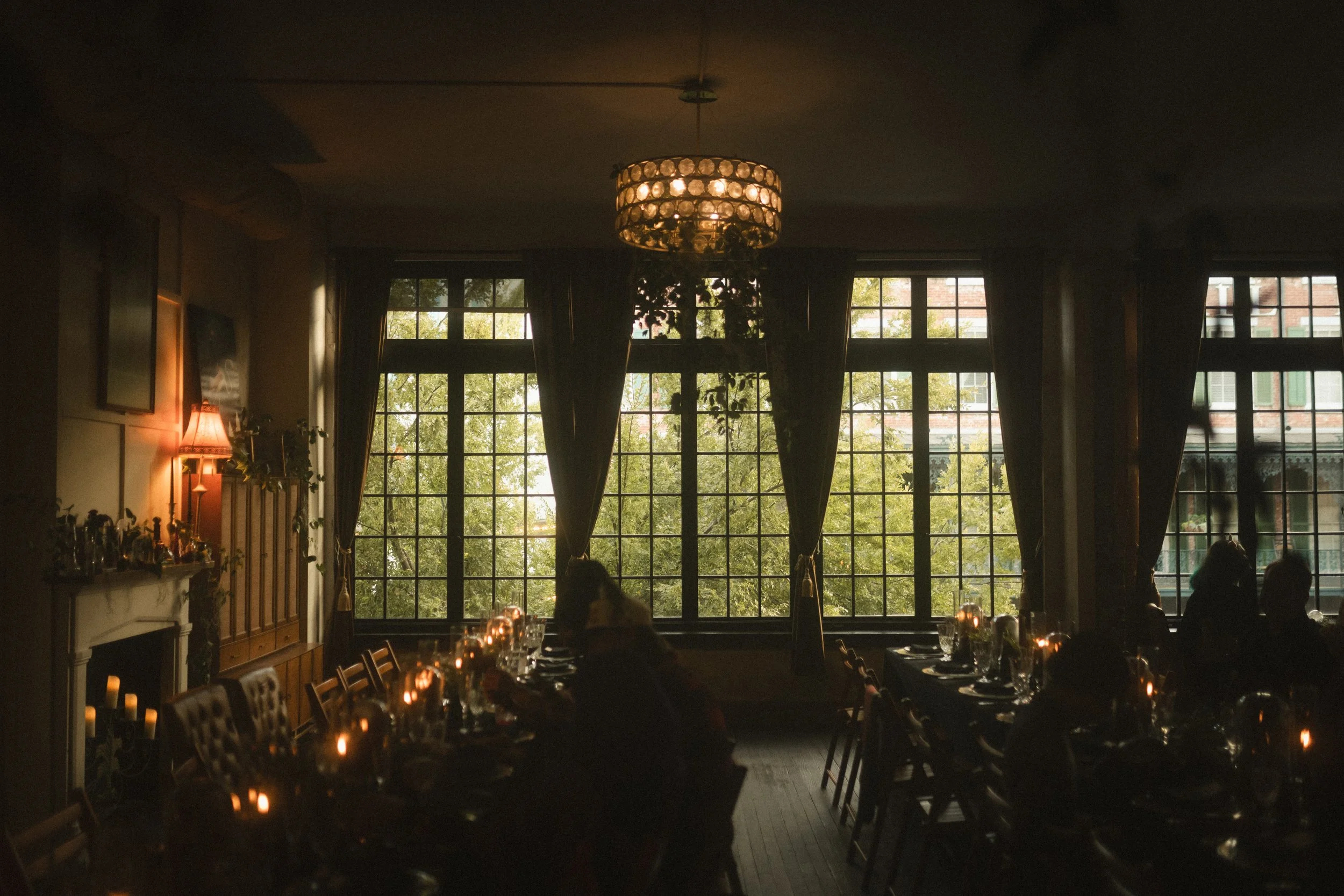 Dimly lit restaurant dining room with candlelit tables, large curtained windows, and a crystal chandelier