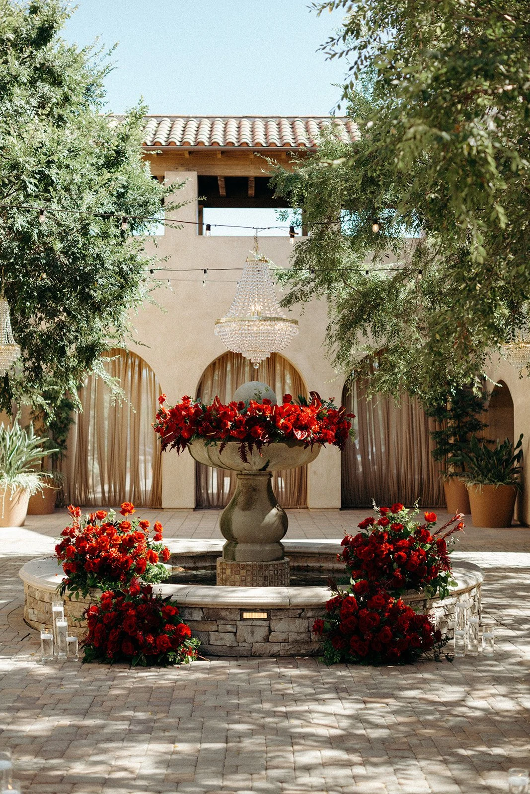 Elegant outdoor courtyard with a fountain decorated with red flowers, surrounded by greenery and large floral arrangements, with chandeliers hanging and soft sunlight.