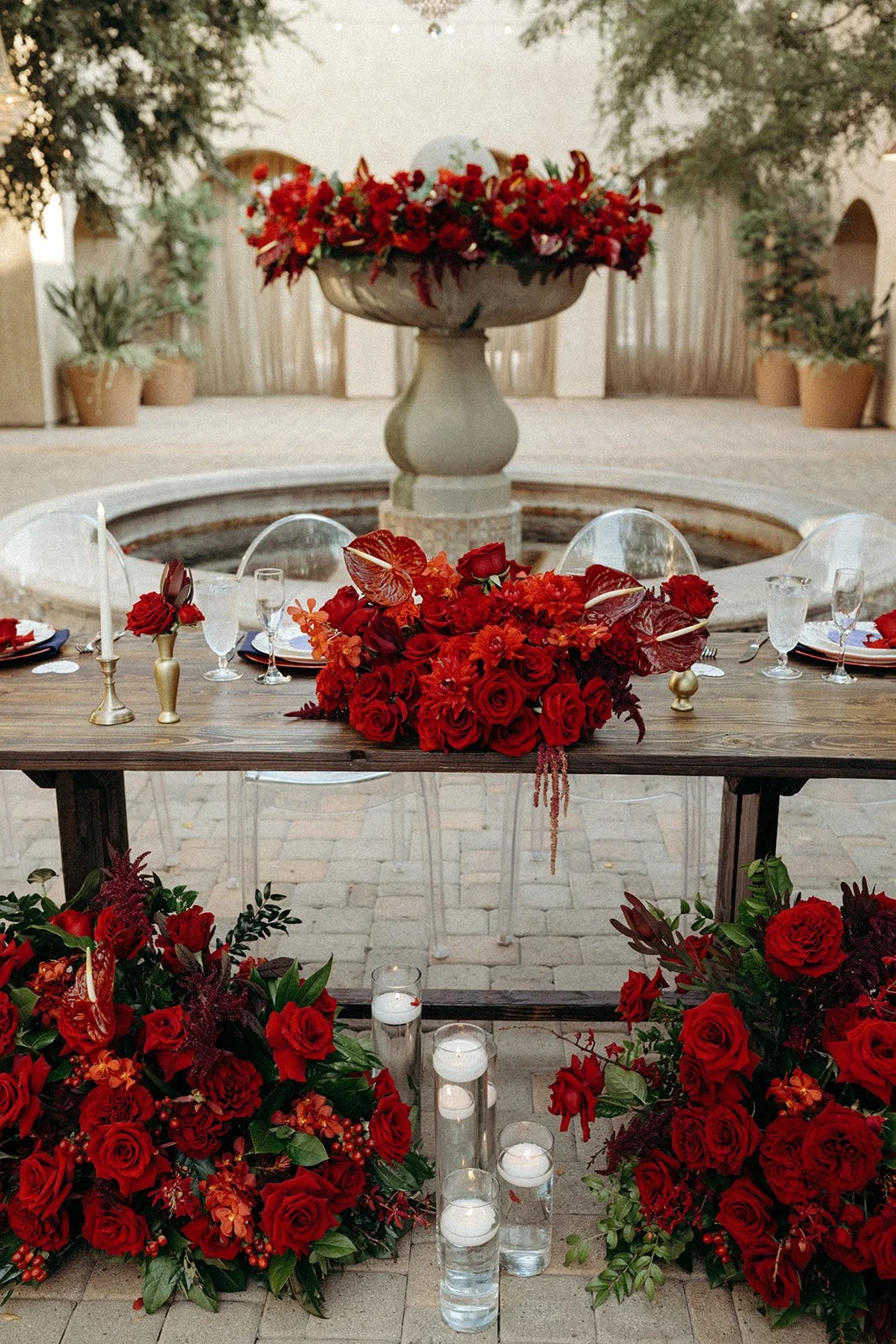 A beautifully decorated outdoor table with red floral arrangements, candles, and glassware, set in front of a fountain with a large floral centerpiece and string lights overhead.
