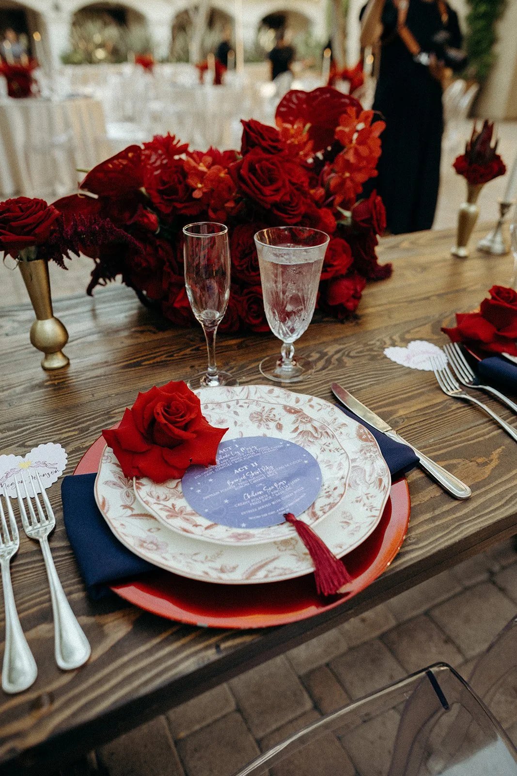 A decorated table at a wedding or special event with a large floral centerpiece of red roses and other red flowers. There are two empty glasses, a floral-patterned plate with a blue and white event menu, a red napkin, and a red rose on the plate. Sil