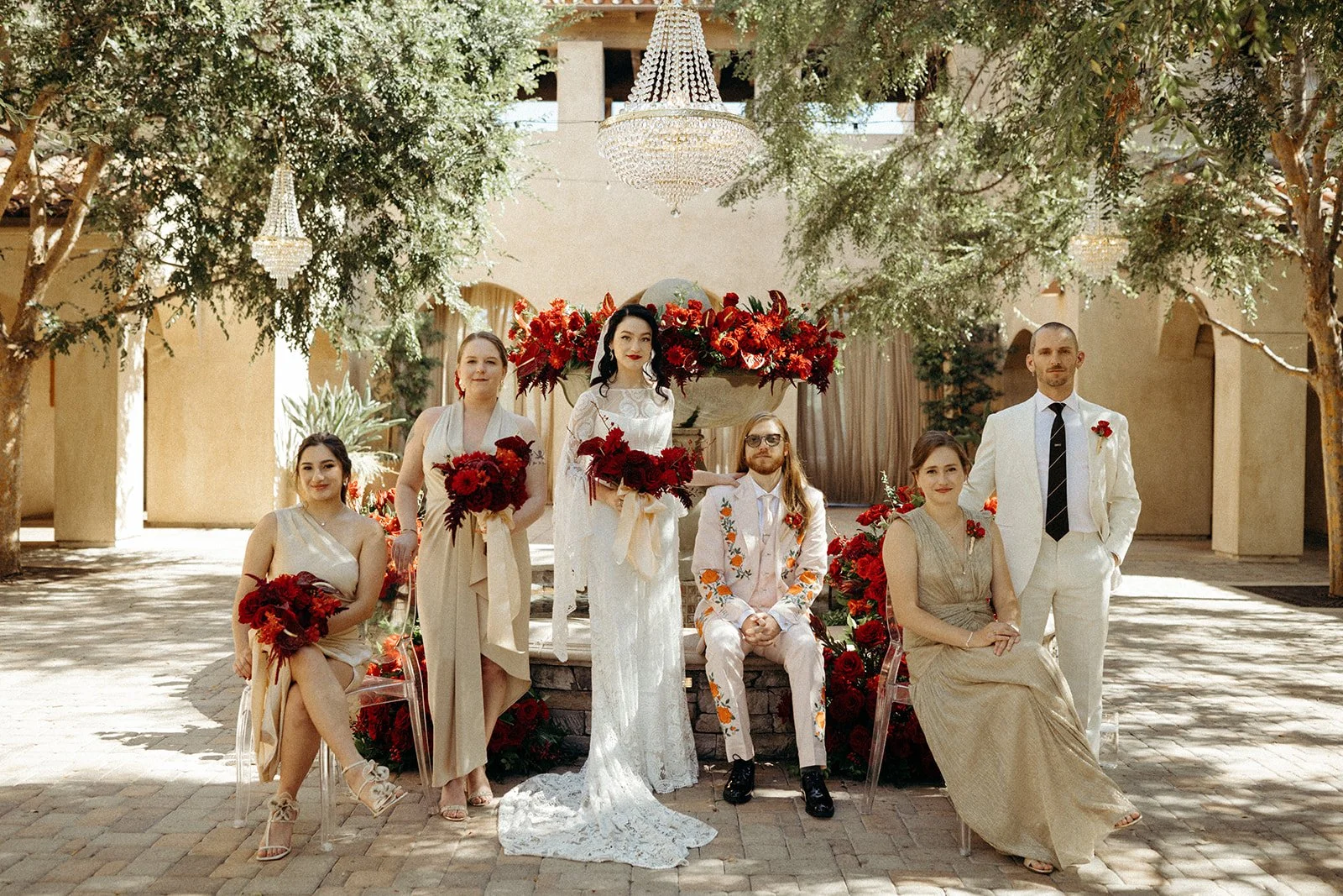A wedding party with seven people, including the bride and groom, standing outdoors under trees and chandeliers, surrounded by red flowers.