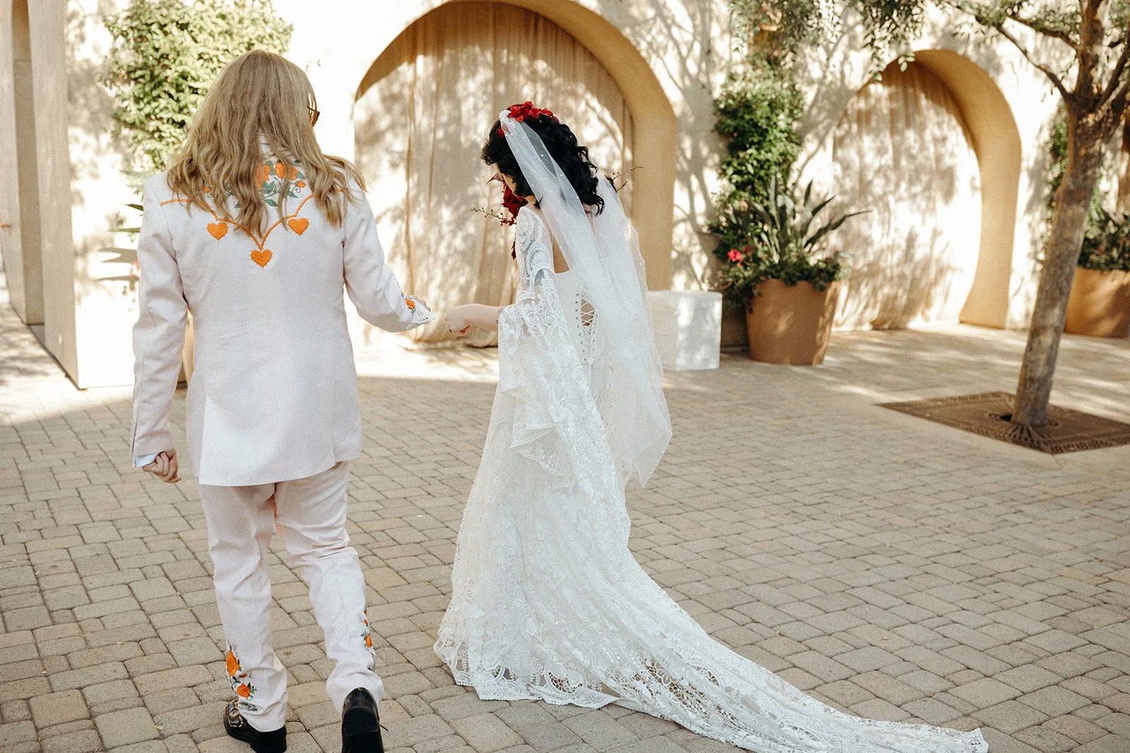 A groom with long blonde hair, wearing a white suit with orange embroidered hearts, walks hand in hand with a bride dressed in a white lace wedding gown and veil, in an outdoor setting with potted plants and arched structures.