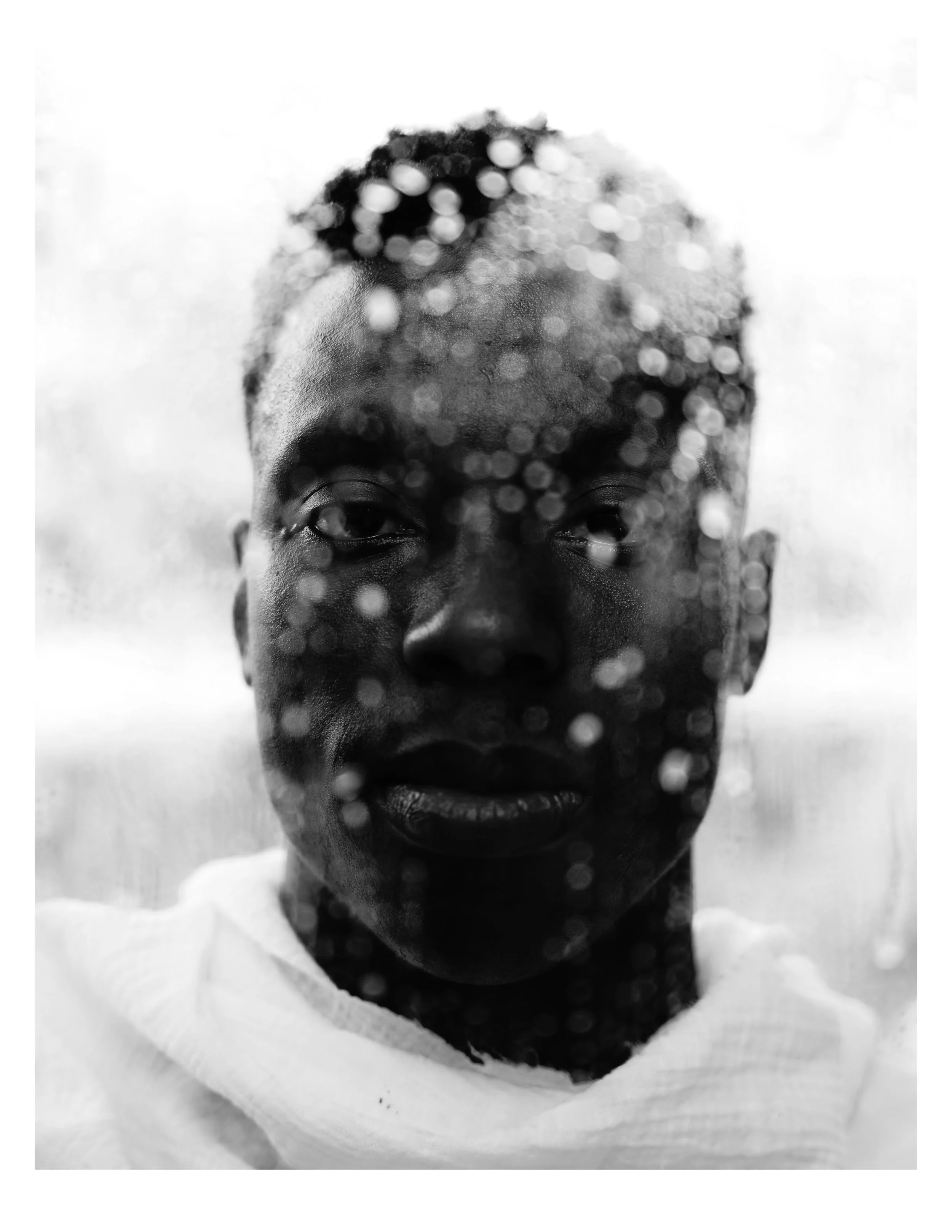Black and white portrait of a person with water droplets on a glass surface in front of their face, looking directly at the camera.