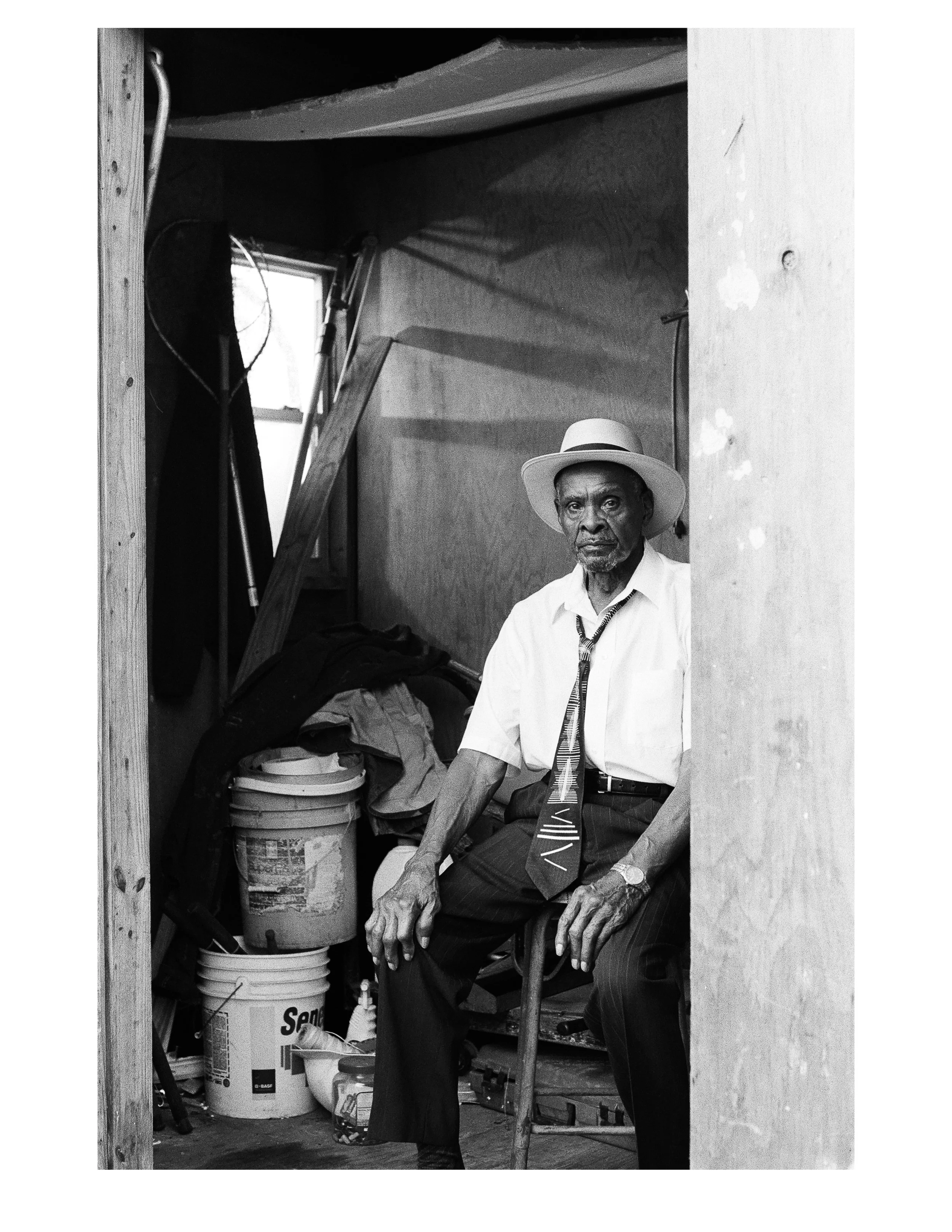 An elderly man wearing a white shirt, patterned tie, watch, and a fedora hat sitting on a wooden chair inside a cluttered room with wooden walls, a small window, and various items like paint buckets, a mop, and clothing.