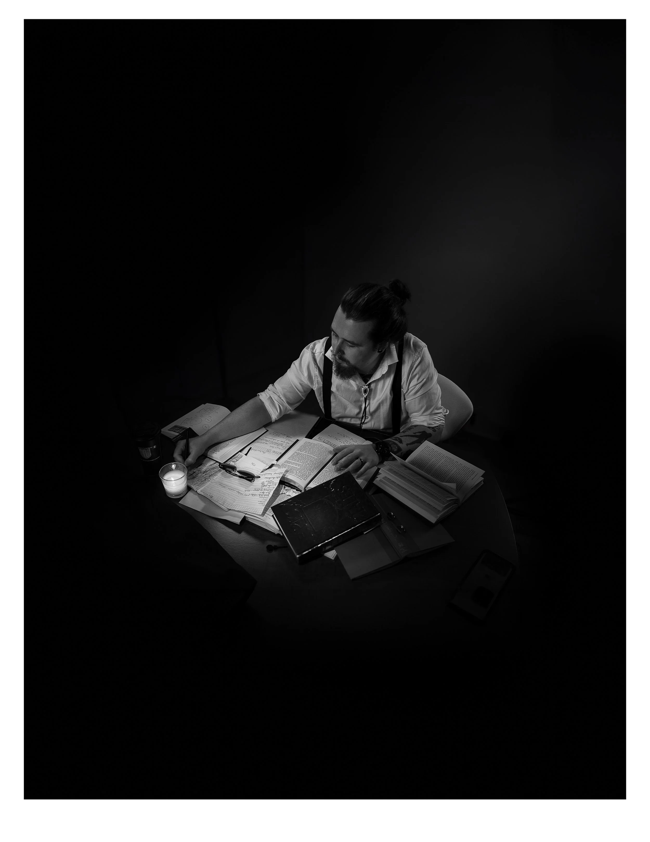 A man with a beard and long hair in a bun, sitting at a cluttered desk with books, papers, a notebook, a pen, a phone, and a candle, working in a dark room.