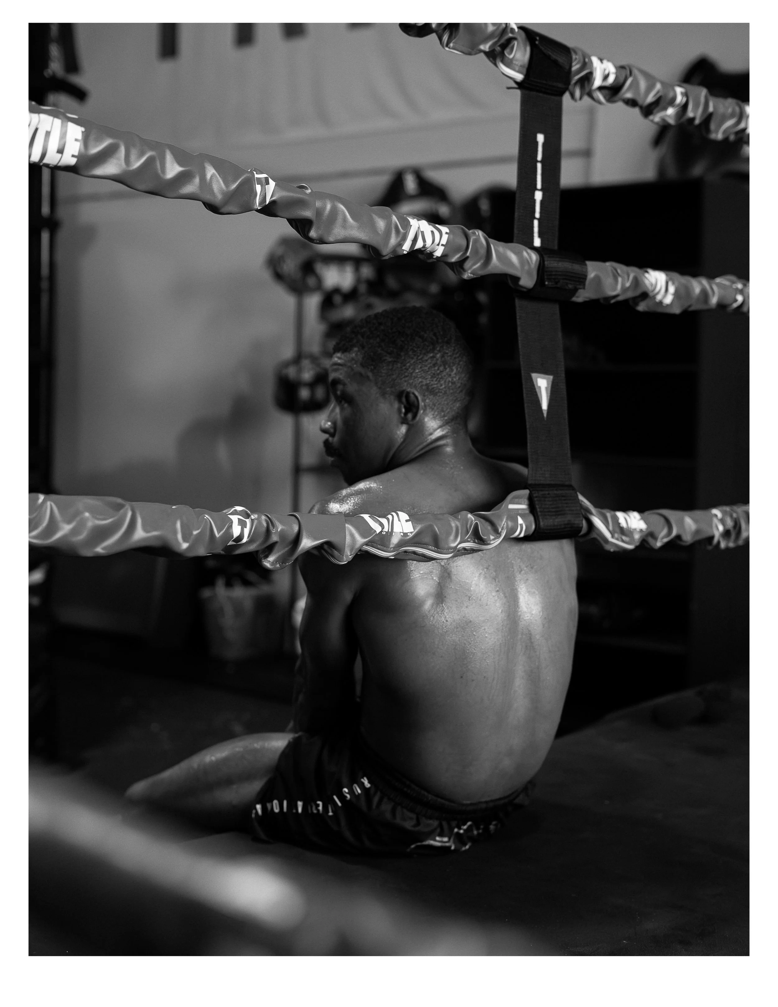 A shirtless young man with a short haircut sitting on a workout mat in a gym, surrounded by boxing or gym equipment, with boxing rings or ropes in the foreground.