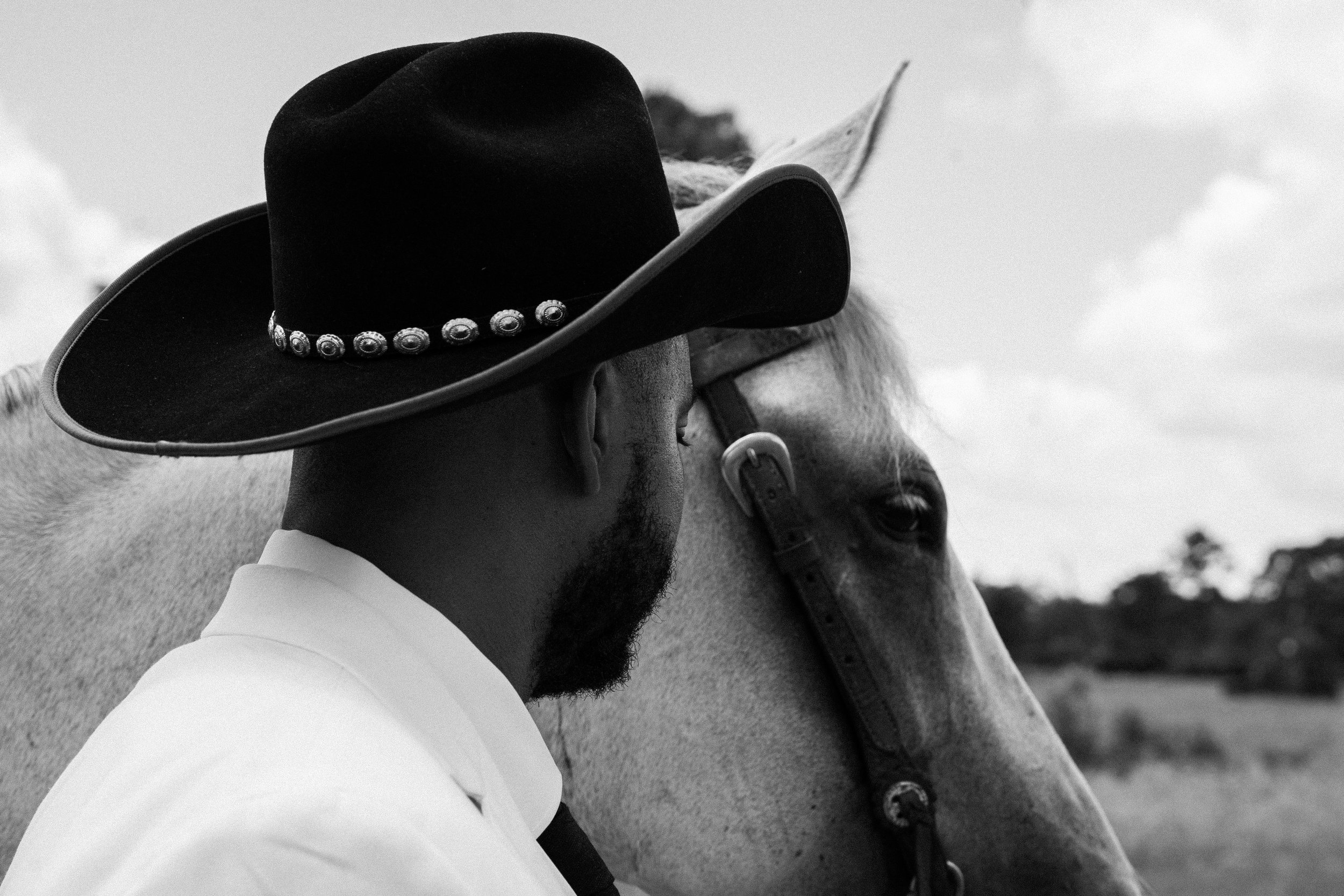 A man in a white shirt and a cowboy hat with decorative band, close to a light-colored horse, in an outdoor setting. Portrait by Marc-Antony Guillory, owner of Zero Sense Productions.