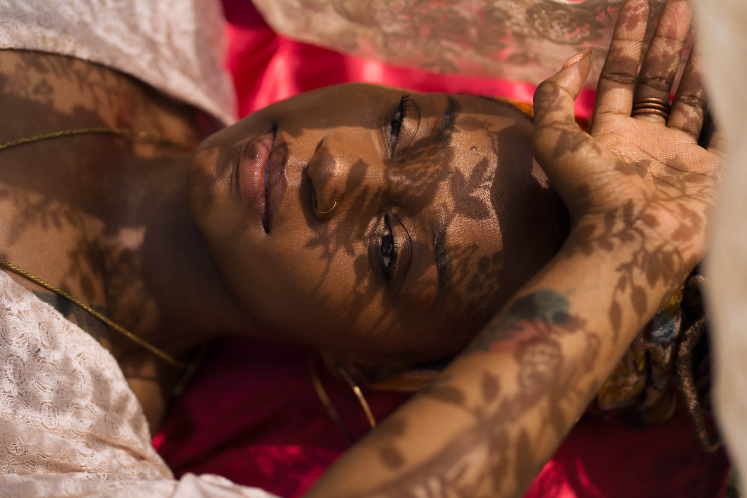 Portrait taken by photographer Marc-Antony Guillory, Zero Sense Productions, of a woman with dark skin lying down, partially covered with a colored cloth, shadows of leaves cast on her face, with tattoos visible on her arm, wearing gold jewelry.