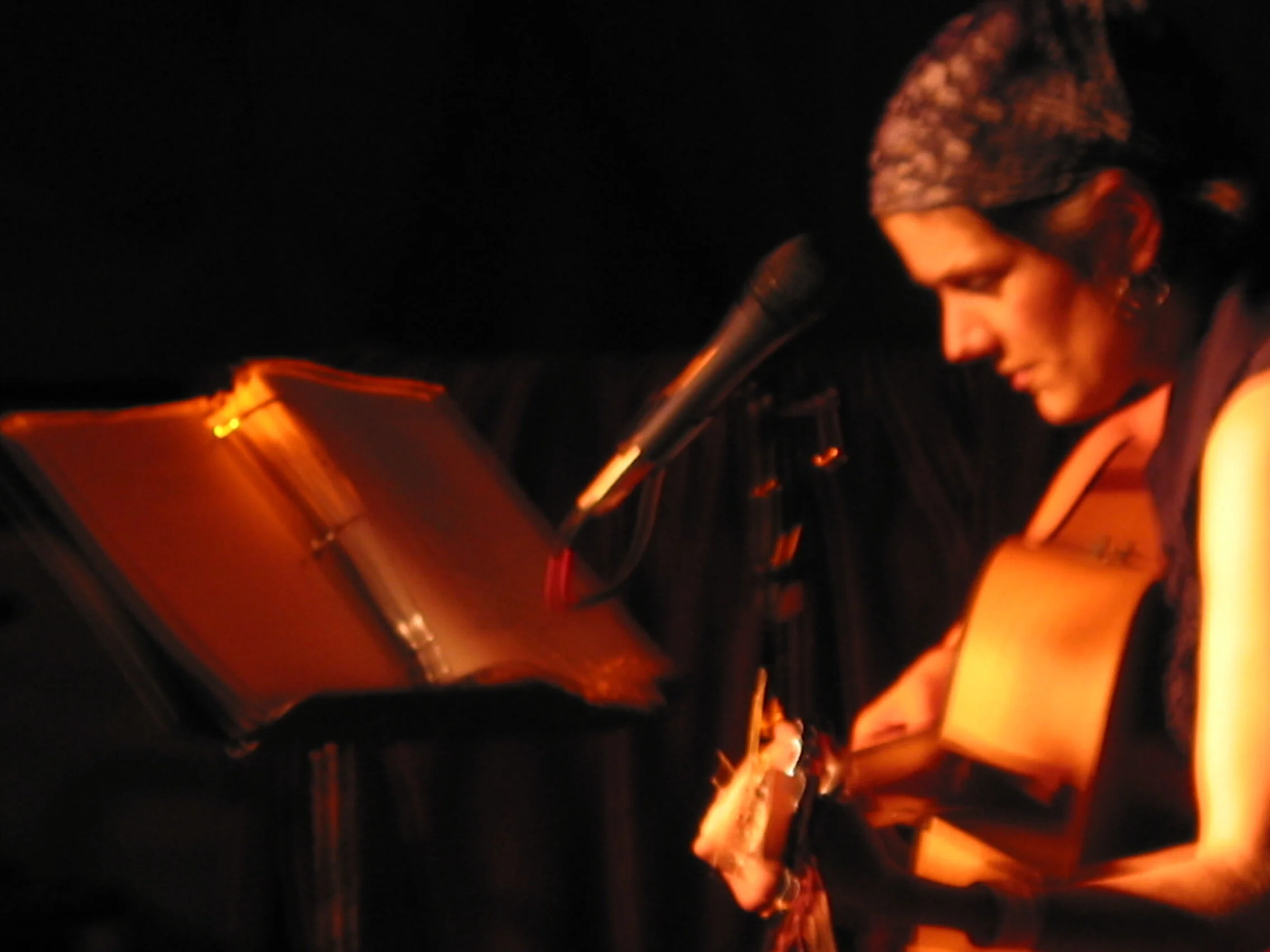 A woman with a headscarf reading from a book at a microphone during a performance.
