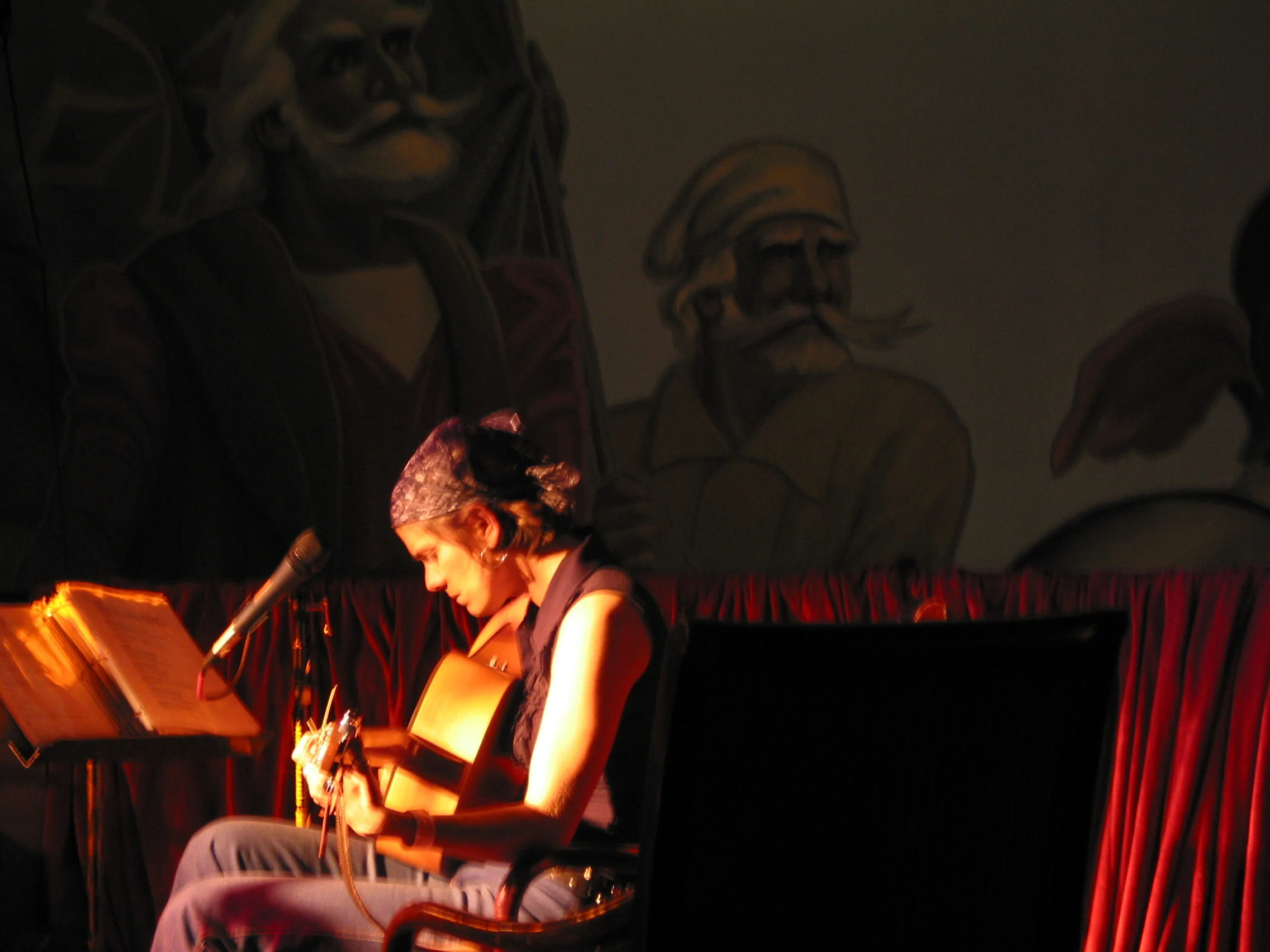 A woman with a bandana playing an acoustic guitar on stage, illuminated by warm lighting, with painted murals of historical figures in the background and red curtains.