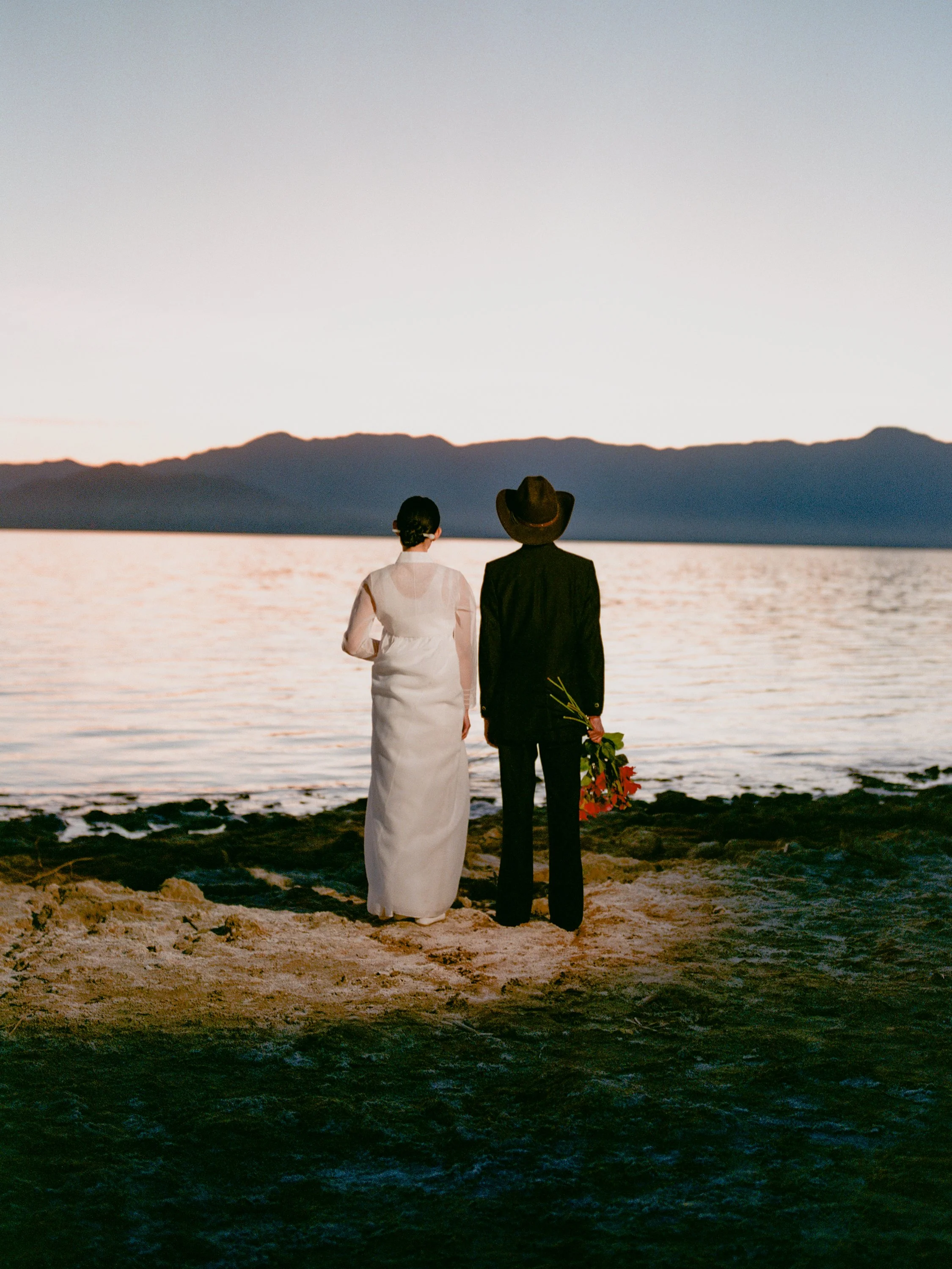 A newly arrived couple looks past at a serene body of water towards the intimidating mountains ahead, as they contemplate where they came from and where their ambitions leads them. This location, Salton Sea, which was once a thriving vacation destina