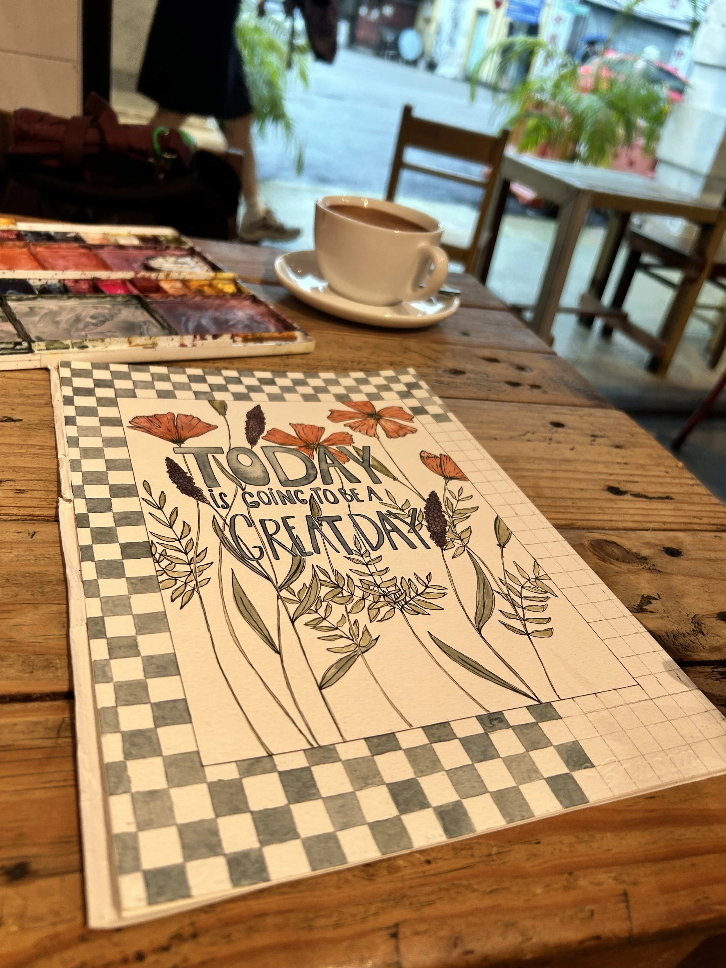 A table in a cafe showcases a colorful motivational sign reading 'Today is going to be a great day' surrounded by illustrated flowers, a cup of coffee, and a watercolor palette set. In the background, there are chairs, a window, and a street scene ou