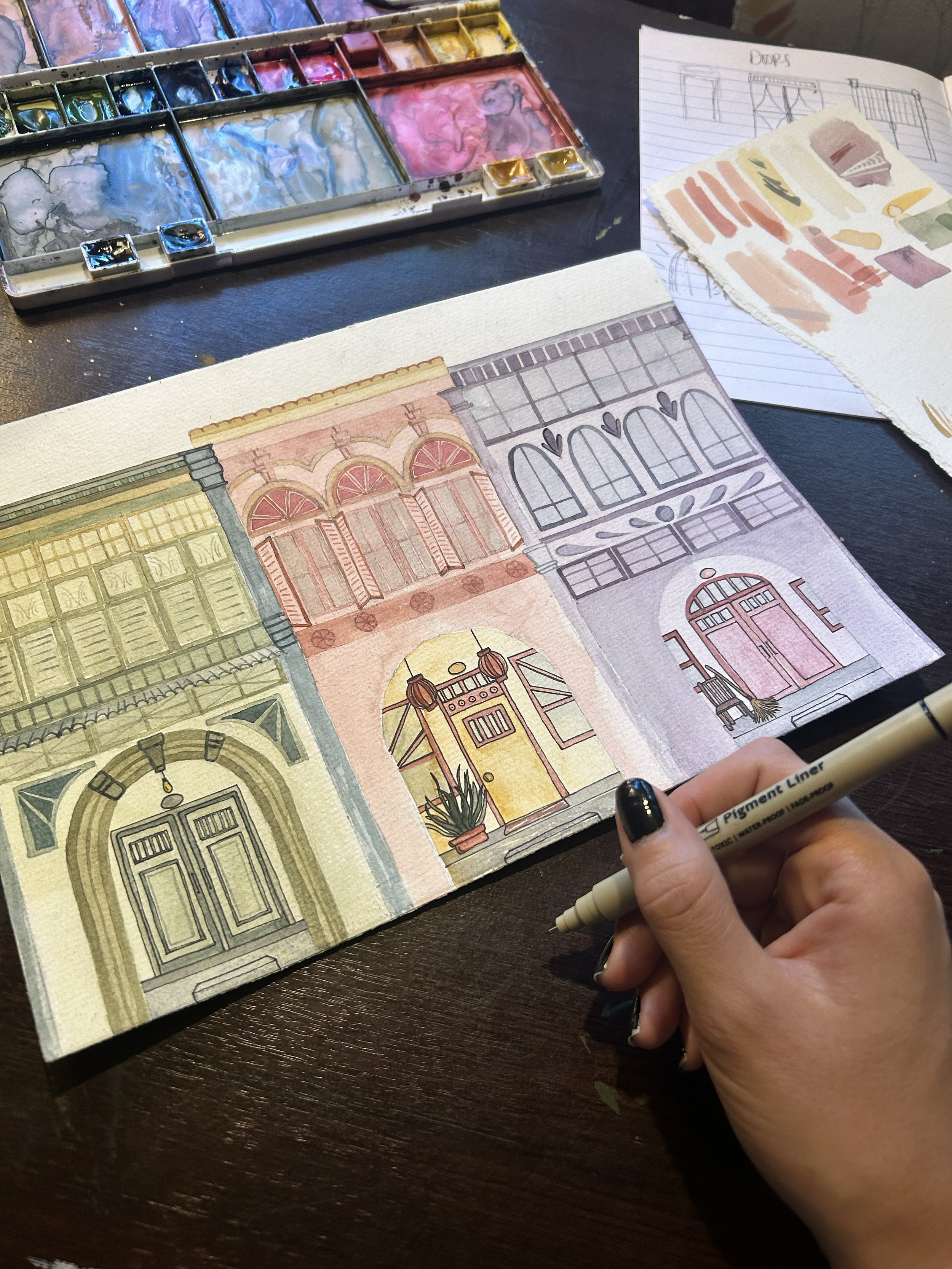 A person is watercolor painting a cityscape of colorful architectural facades on paper, with watercolor paints and sketches visible on the desk.