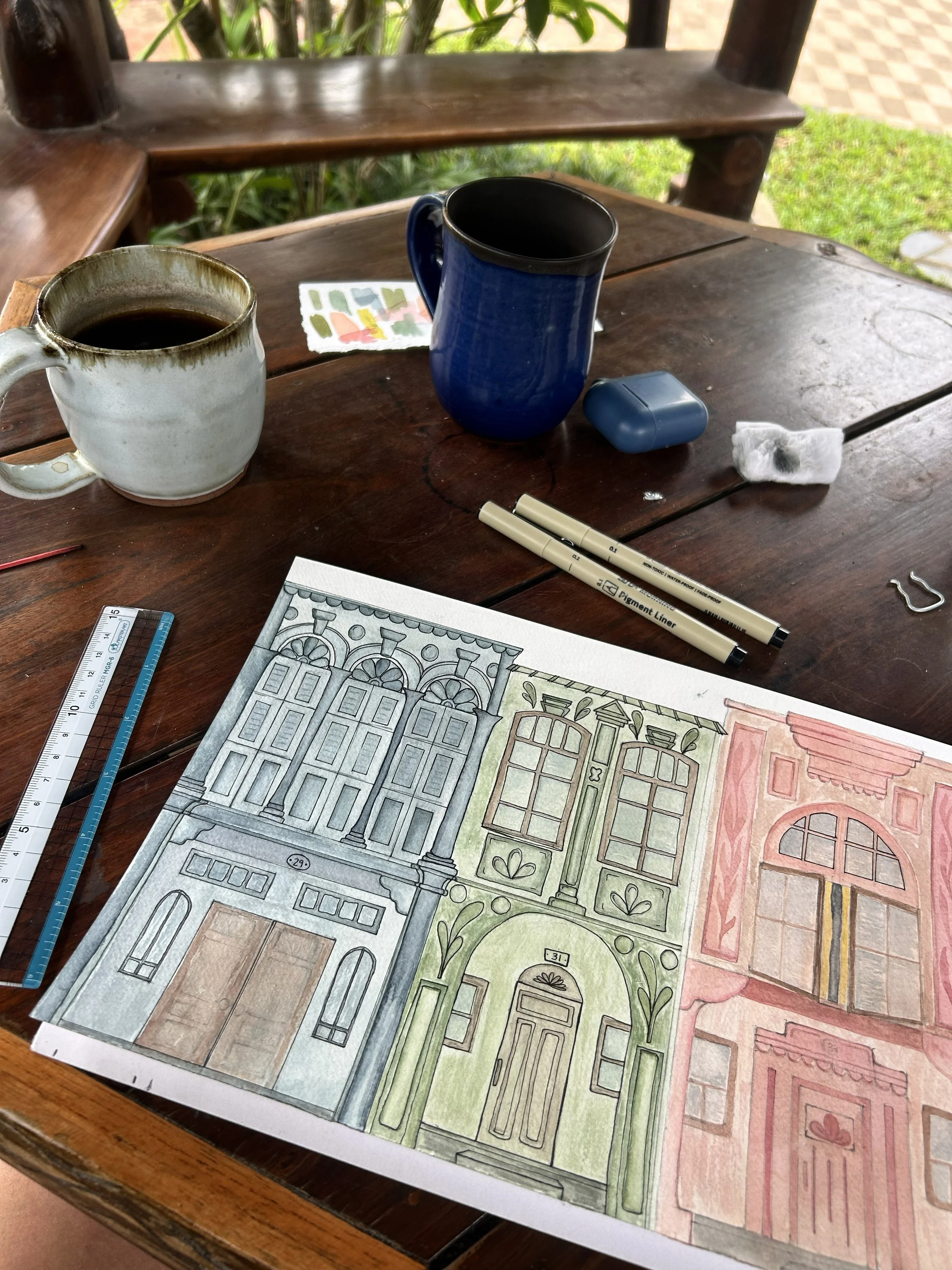 Colored architectural sketches of three different building facades on a table, surrounded by drawing tools, coffee mugs, and other small items.