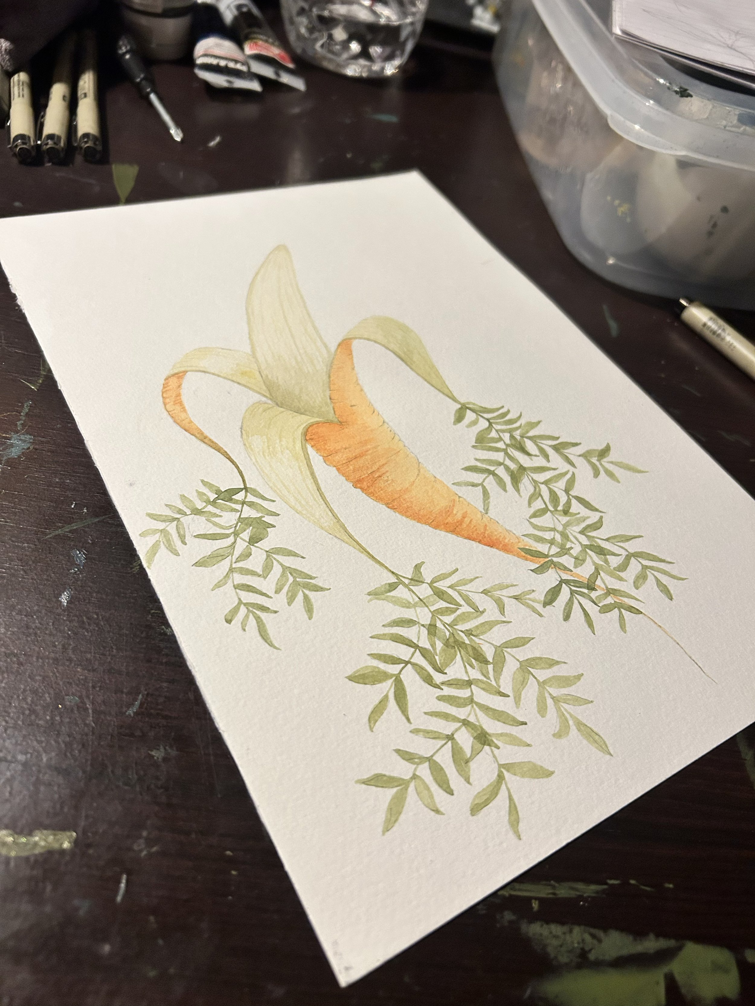 A watercolor painting of a carrot with green leaves on a white sheet of paper, placed on a dark wooden table surrounded by art supplies.