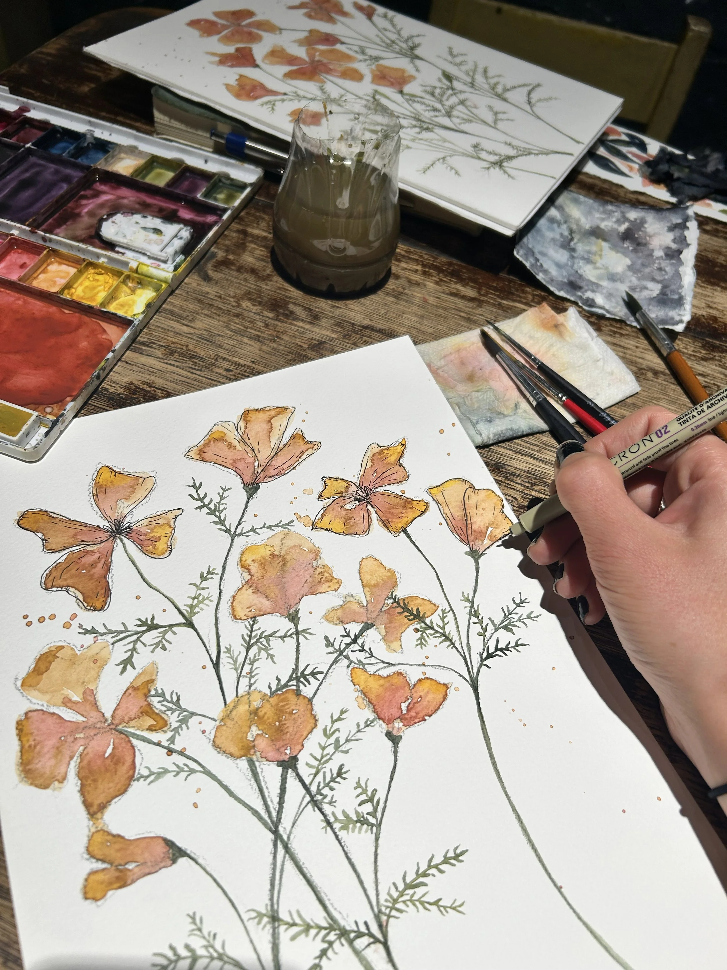 Watercolor painting of orange and pink flowers with green stems and leaves on white paper, surrounded by watercolor paints, brushes, and a glass of water on a wooden table.