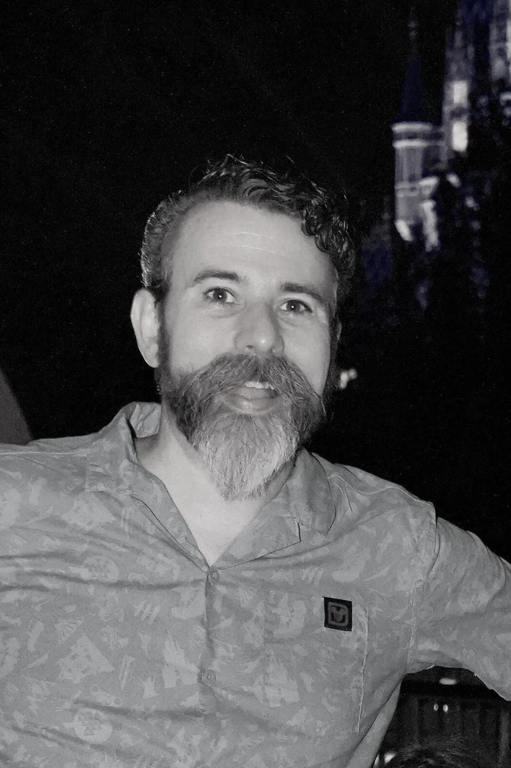 Black and white image of a 40-something year old white man with short, dark, curly hair and a mixed black and white beard that comes to a point. The man also sports a glorious mustache.