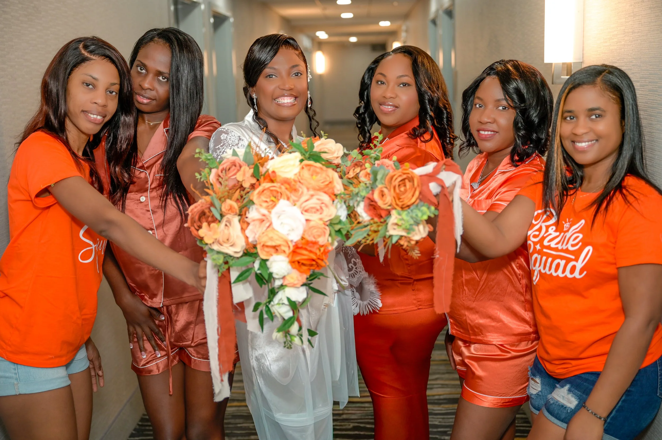 Group of women celebrating an event, holding a flower bouquet, dressed in casual and satin sleepwear, in a hallway.