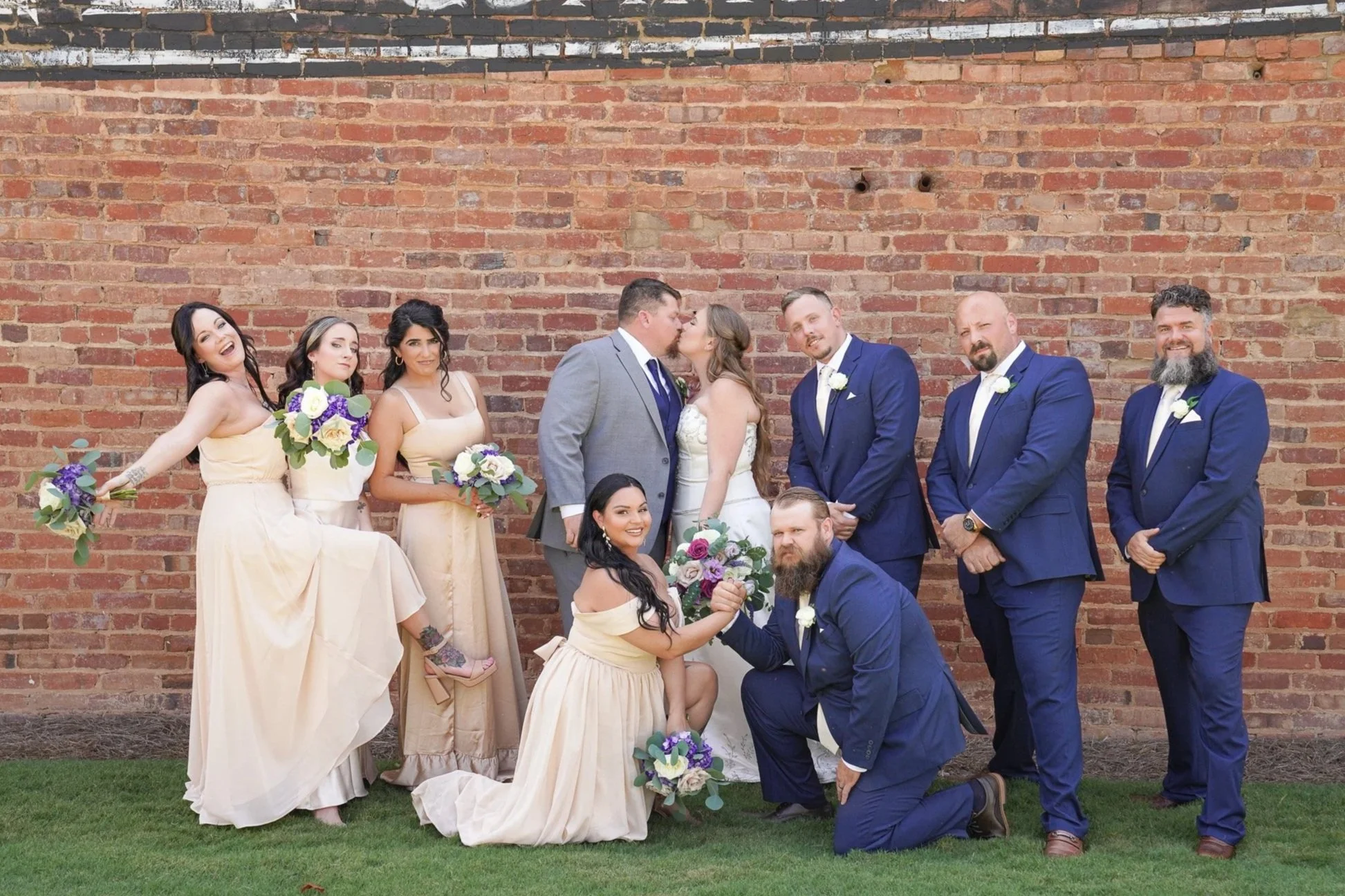 A wedding party standing and kneeling in front of a brick wall, with the bride and groom kissing in the center. The bride is wearing a white strapless gown, and the groom is in a gray suit. Bridesmaids are in peach dresses holding purple and white bouquets, and groomsmen are in navy blue suits with white shirts and ties, some with white boutonnieres. A woman in a peach dress kneels in front holding a bouquet, and a man in a navy suit kneels beside her. The group is smiling and posing for the photo.