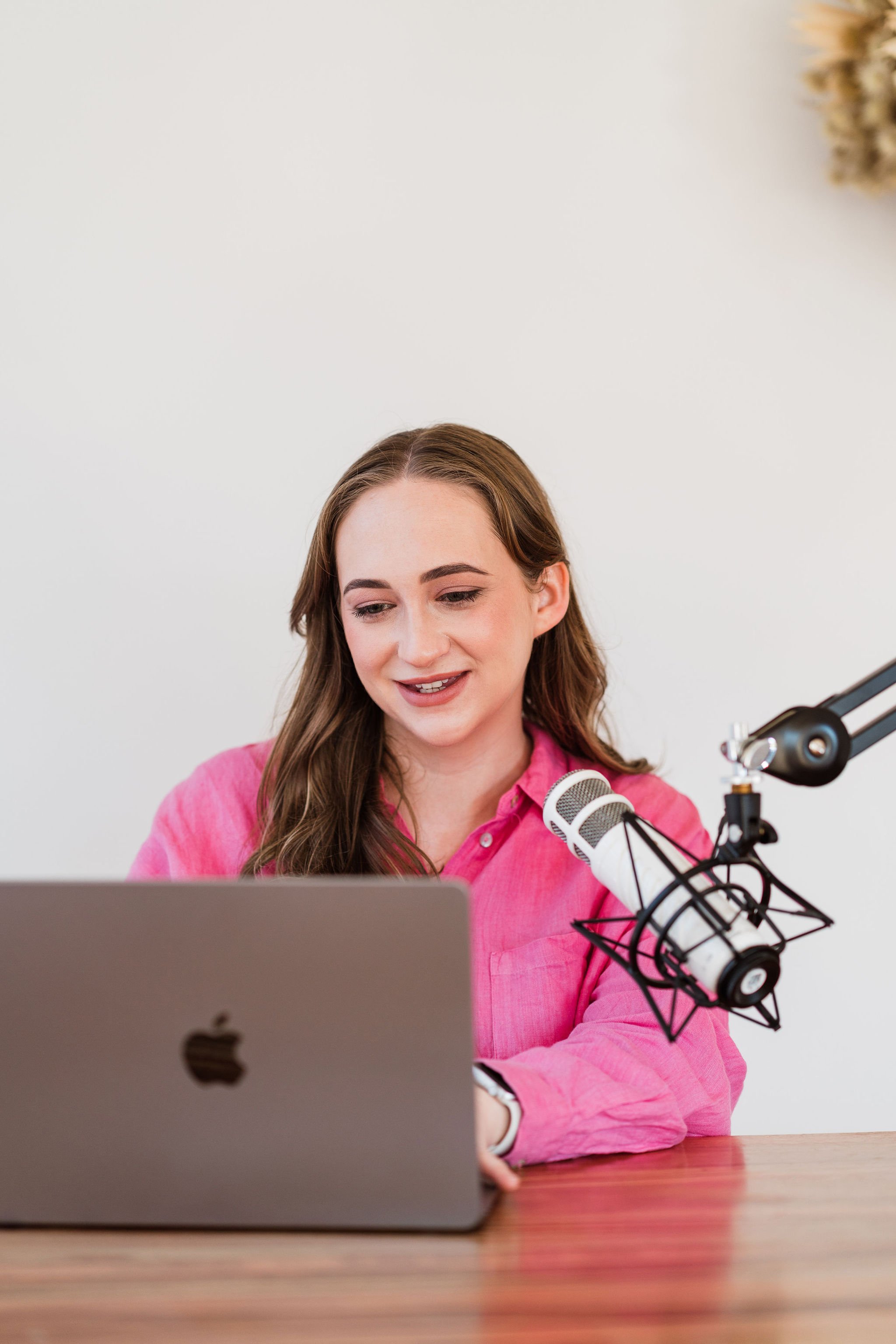 Maxene London sitting at a desk with her laptop open and rode microphone pointed towards her. She is where a pink shirt