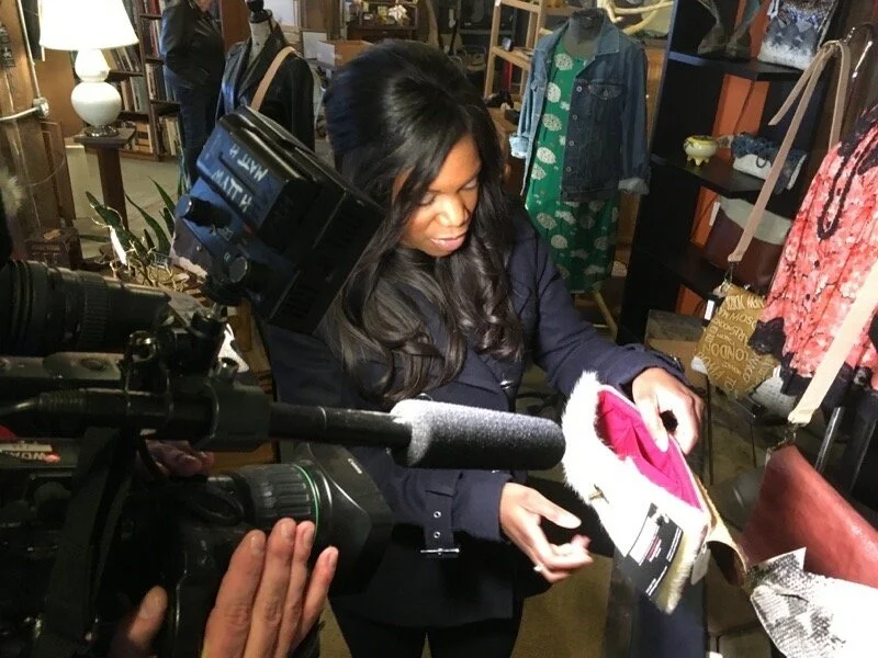 A woman examines a small bag in a store, surrounded by clothing items and accessories. A camera crew is filming her.