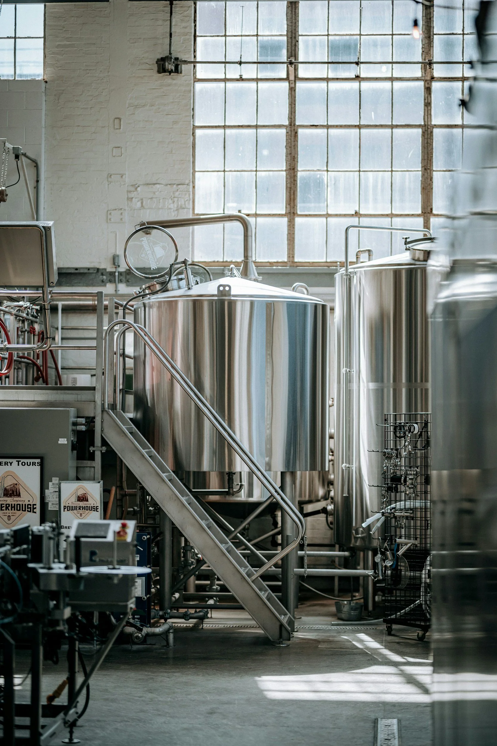 Interior view of a brewery or industrial brewing facility with stainless steel tanks and pipes, large windows letting in natural light.